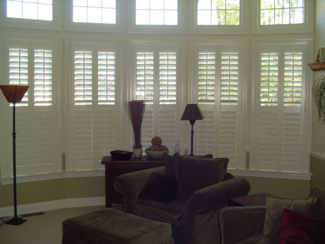 White shutters cover a bay window in a living room; a sofa and a lamp are visible.