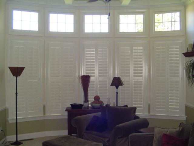 Living room with white shutters on bay windows; brown furniture, floor lamp, and side table.