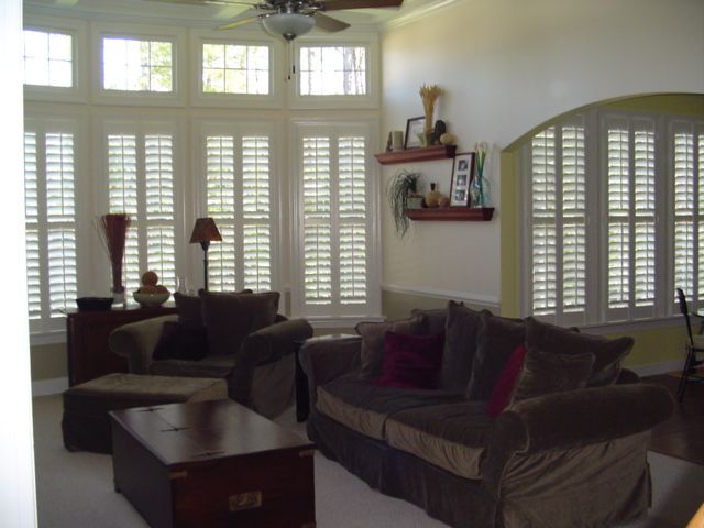 Living room with white shutters, brown furniture, and two shelves with decor.