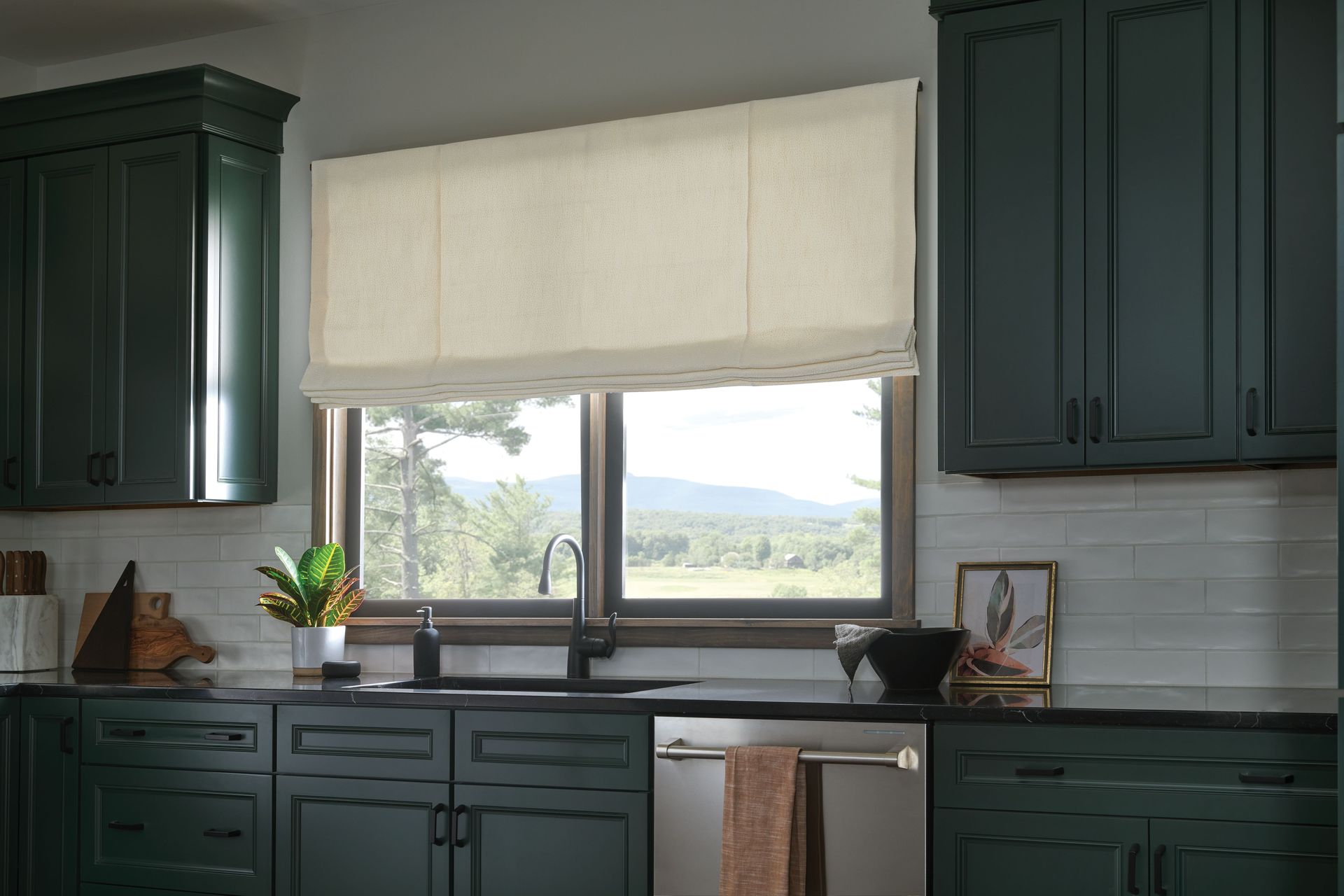 Kitchen with dark green cabinets, a window with a beige Roman shade, and a view of a landscape.