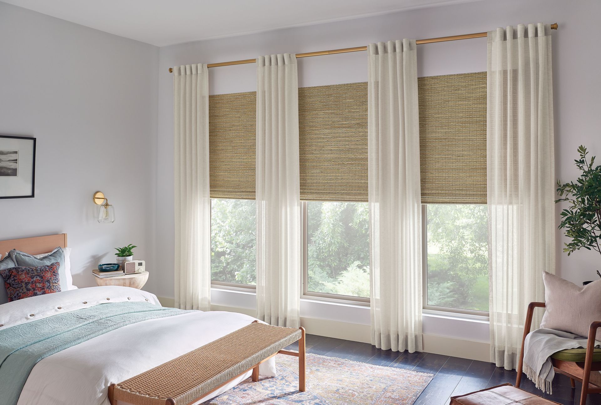 Bedroom with three windows, each with woven blinds and sheer curtains. Includes a bed, bench, and chair.