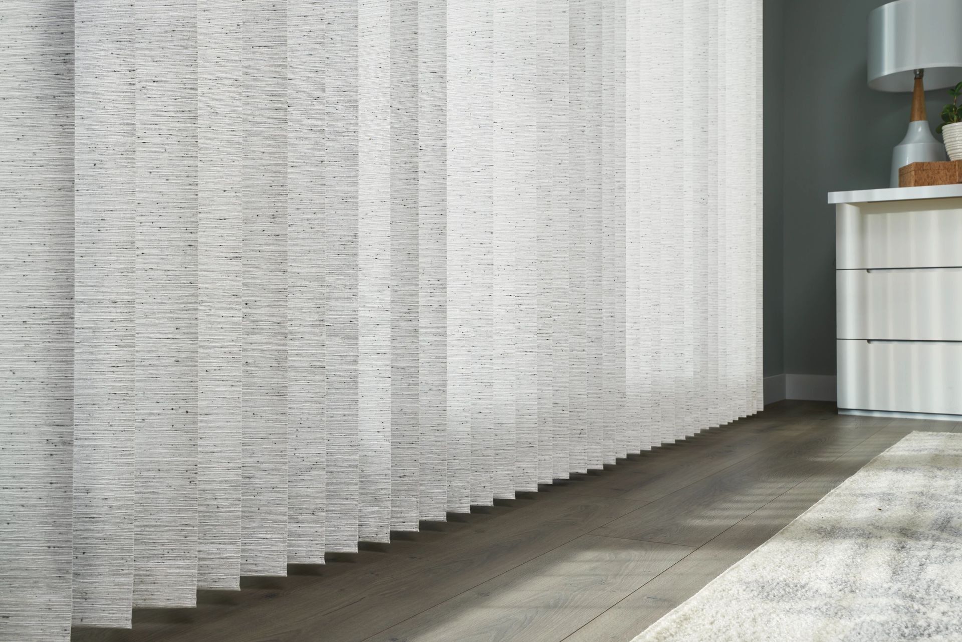 Vertical blinds, gray and textured, partially cover a window in a room with a side table and rug.