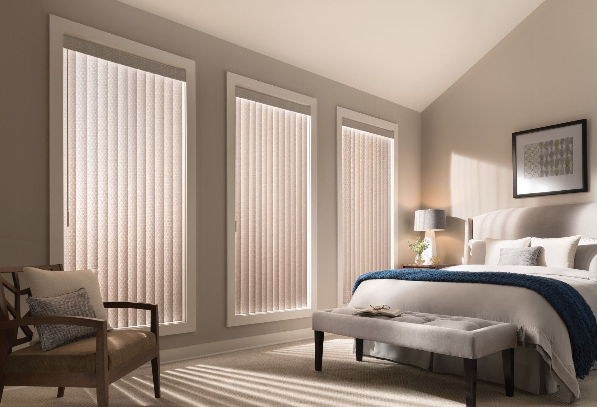 Bedroom with three vertical blinds, bed, bench, chair, and neutral tones.