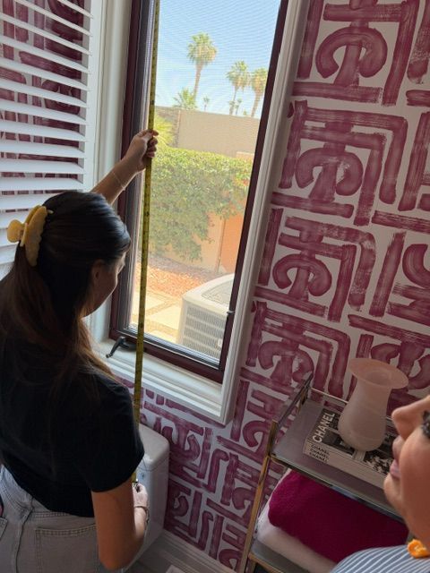 Person measures a window with a yellow tape measure beside pink patterned wallpaper.