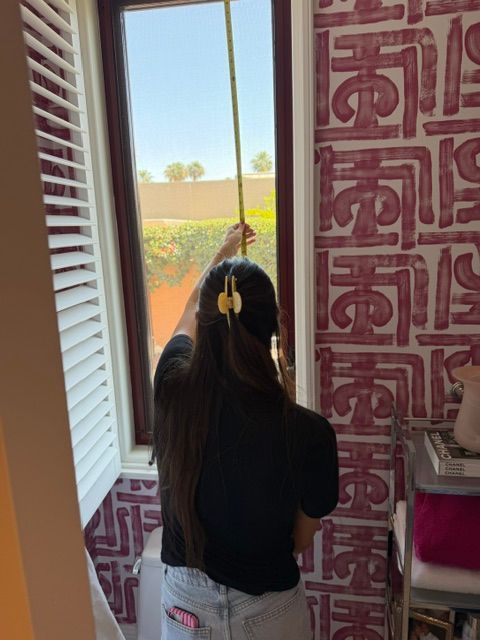 Person standing by a window, looking outside at a bright garden and holding the window frame.