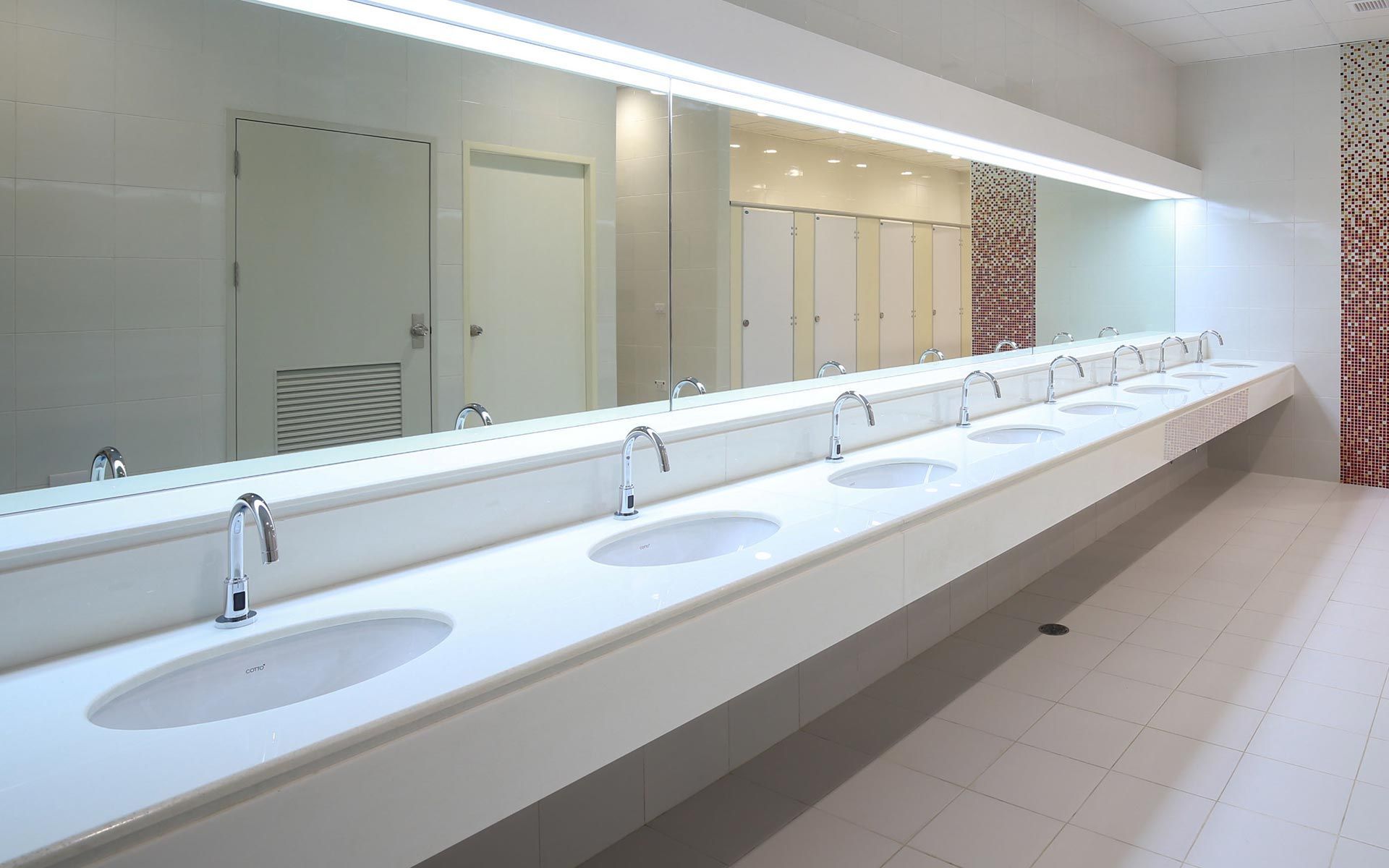Bright public restroom with long white sink counter, mirrors, and stalls in a clean tiled interior.