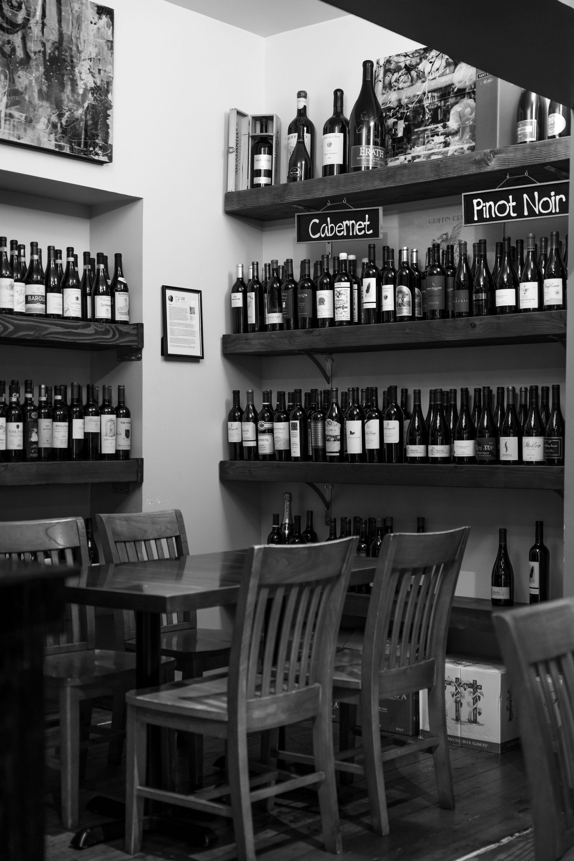 Wine bottles are displayed on shelves in a restaurant, and a table with chairs is in front.