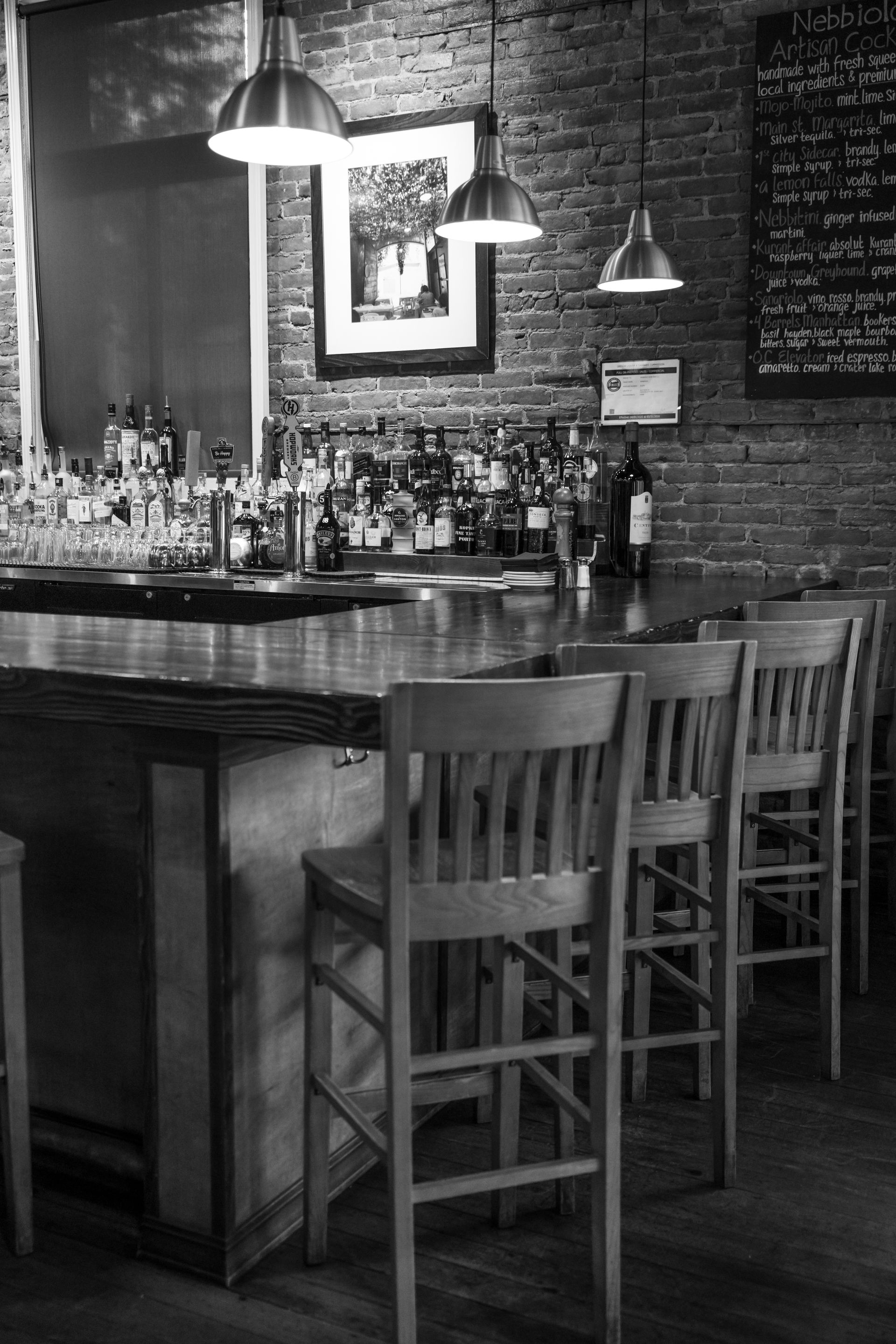 Black and white photo of a bar interior with stools, liquor bottles, and hanging lights. Brick wall in the background.