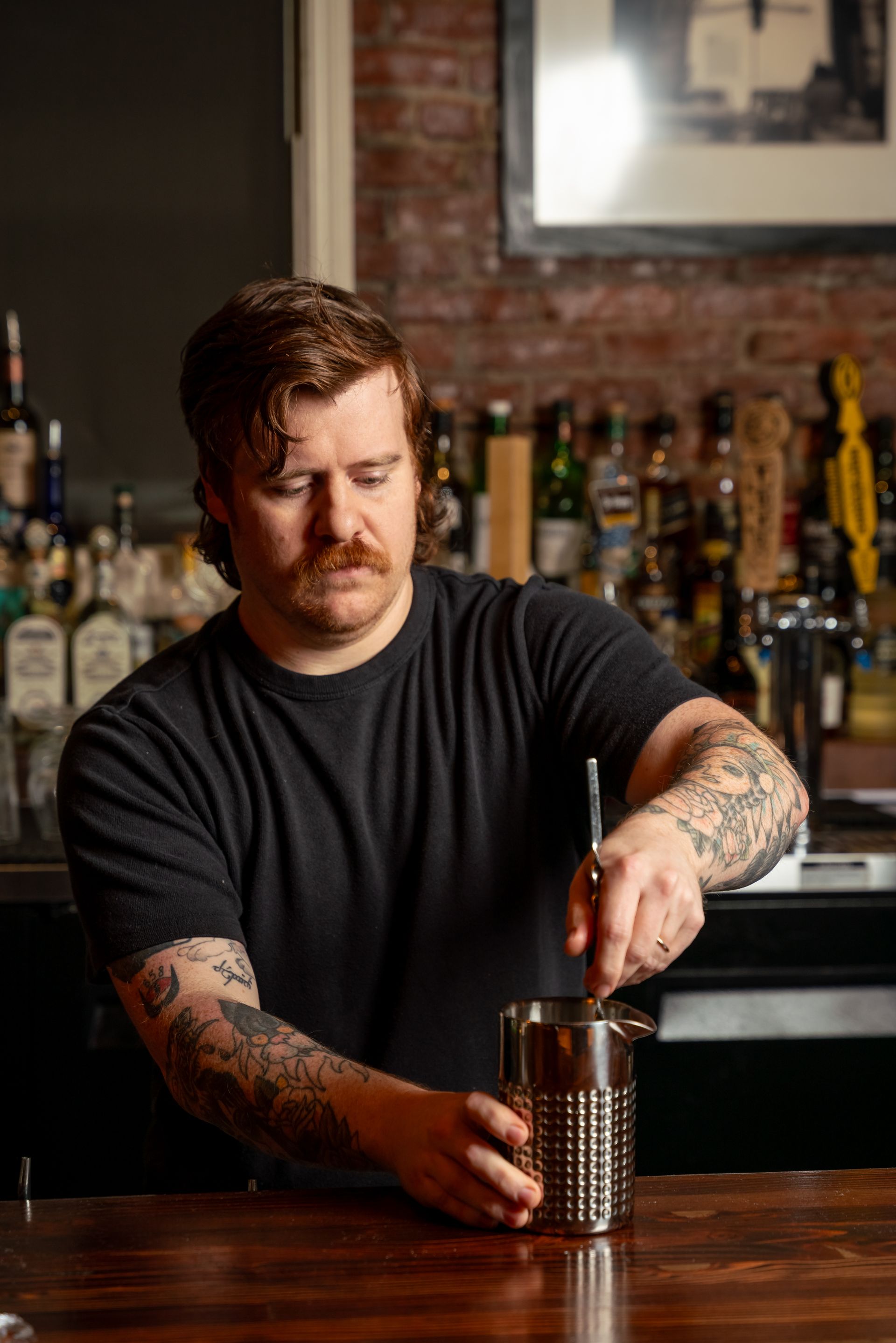 Bartender stirring a drink in a metal shaker behind a bar.