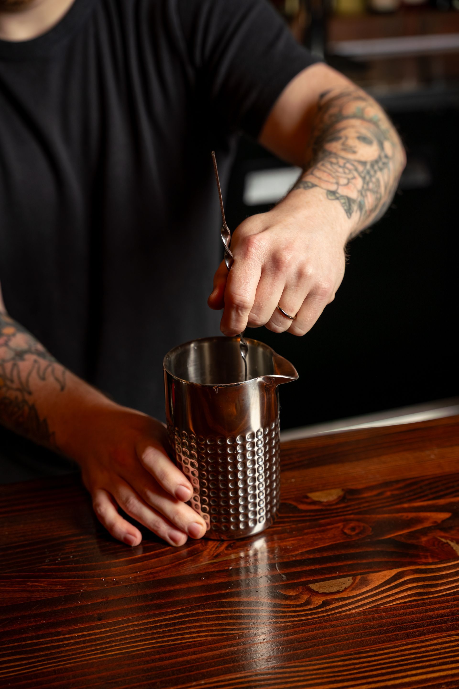 Bartender stirs a drink in a metal shaker on a wooden bar, arms covered in tattoos.
