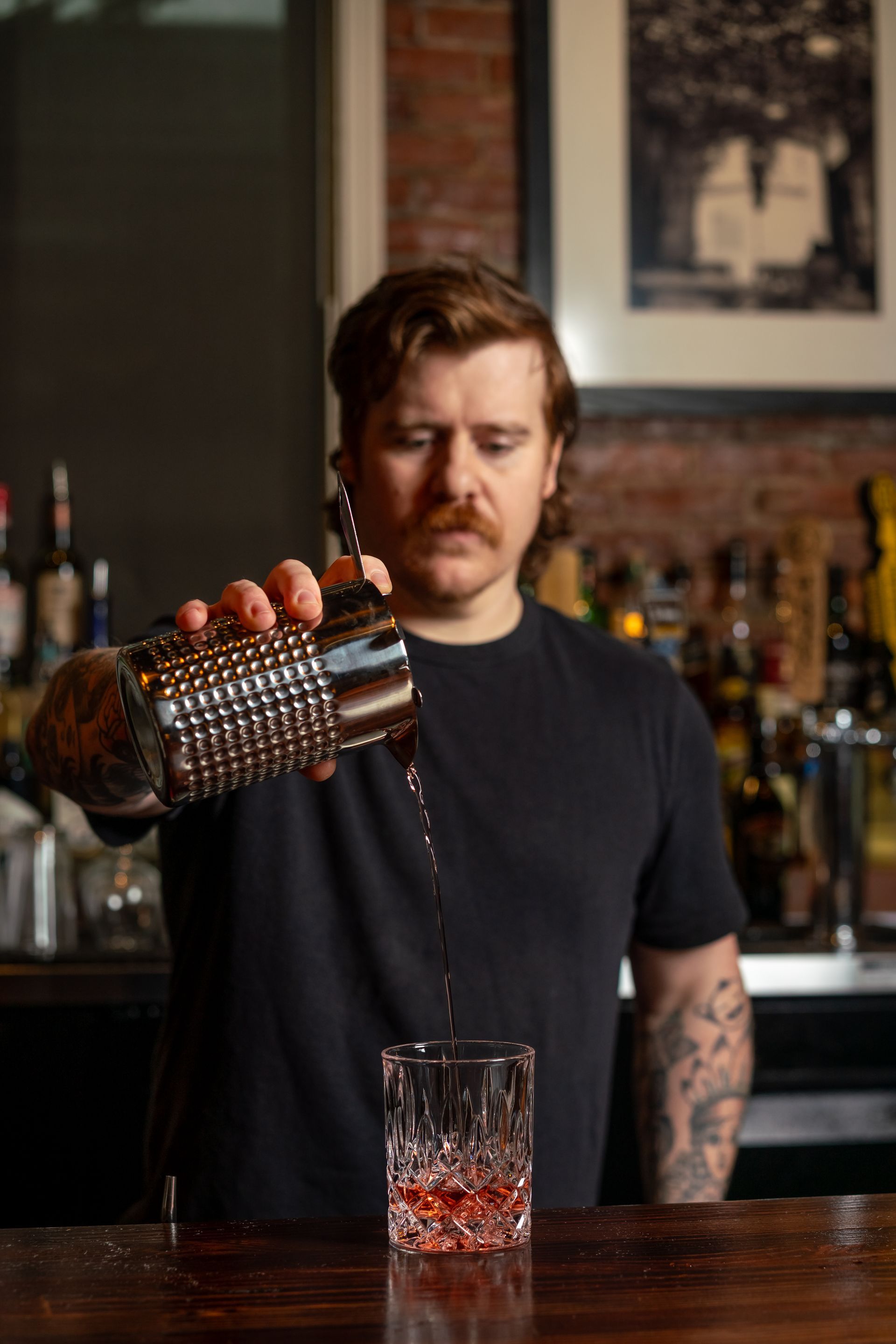 A bartender pours a cocktail from a shaker into a glass at a bar.