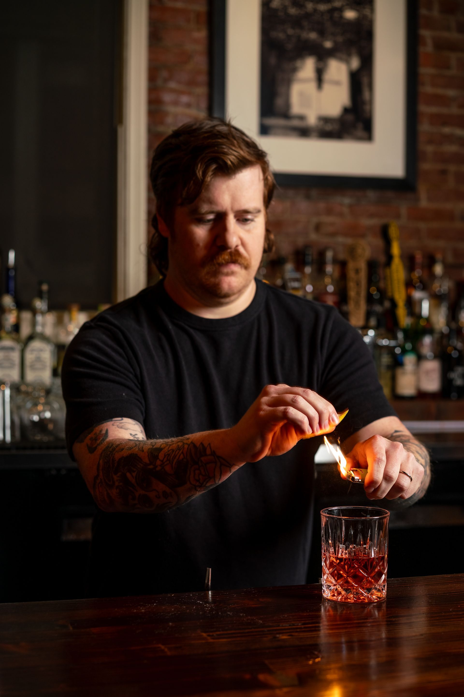 A bartender flames an orange peel over a cocktail in a lowball glass at a bar.