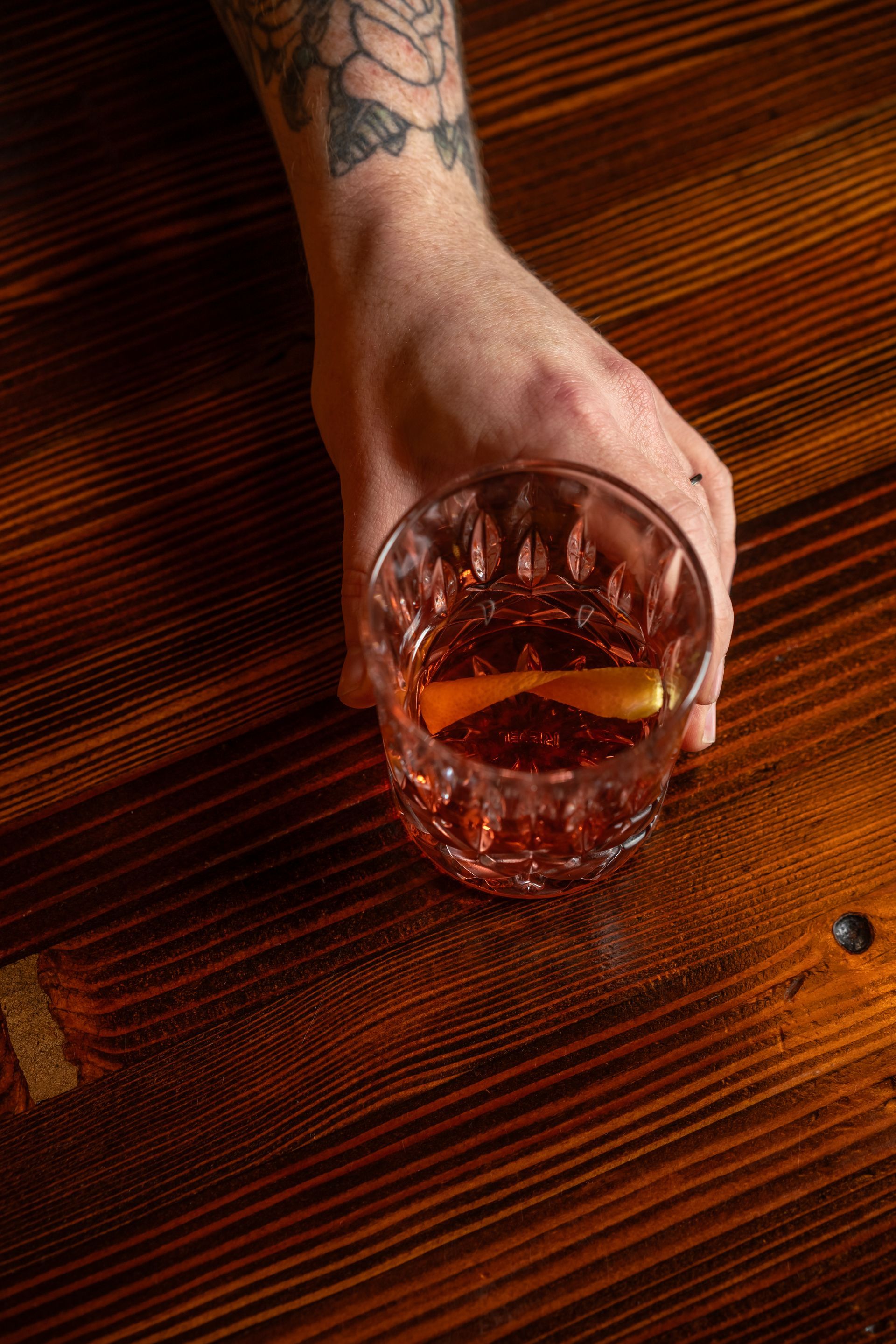 Hand holding a cocktail with an orange peel on a wooden table. Tattoo visible on arm.