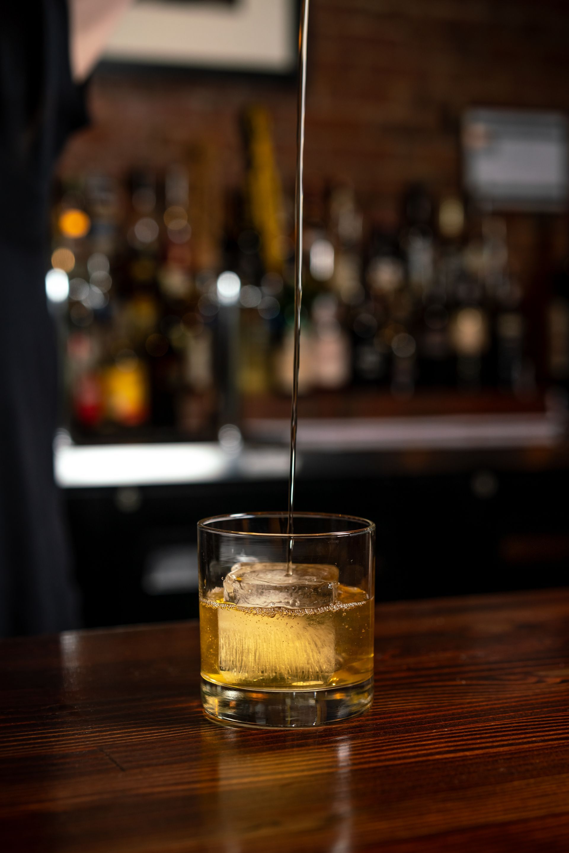 Whiskey is being poured into a rocks glass with a large ice cube on a wooden bar, with a blurred bar backdrop.