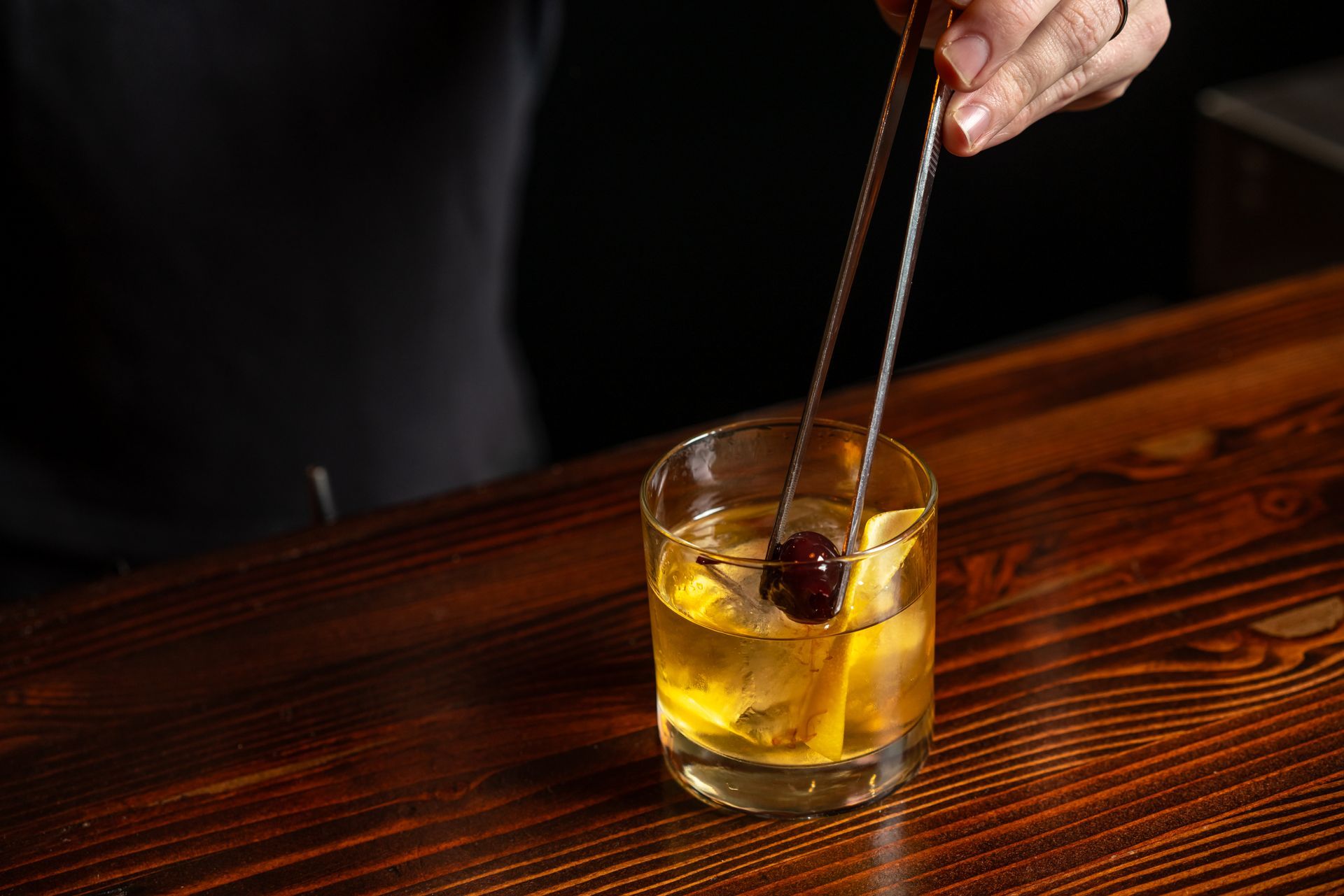 Bartender garnishes an Old Fashioned cocktail with a cherry using tongs on a wooden bar.