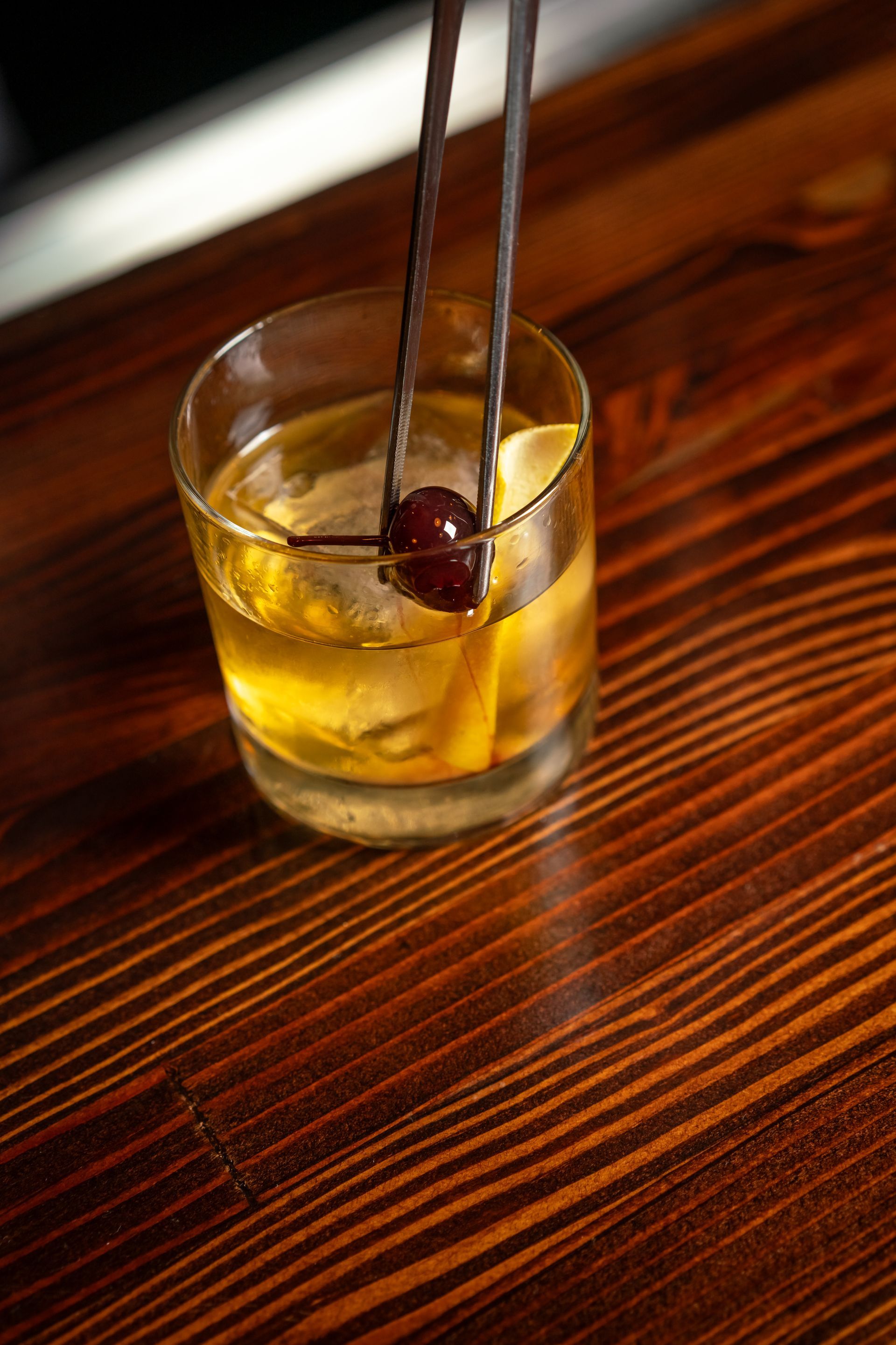 Cocktail being garnished with a cherry using tongs on a wooden bar.