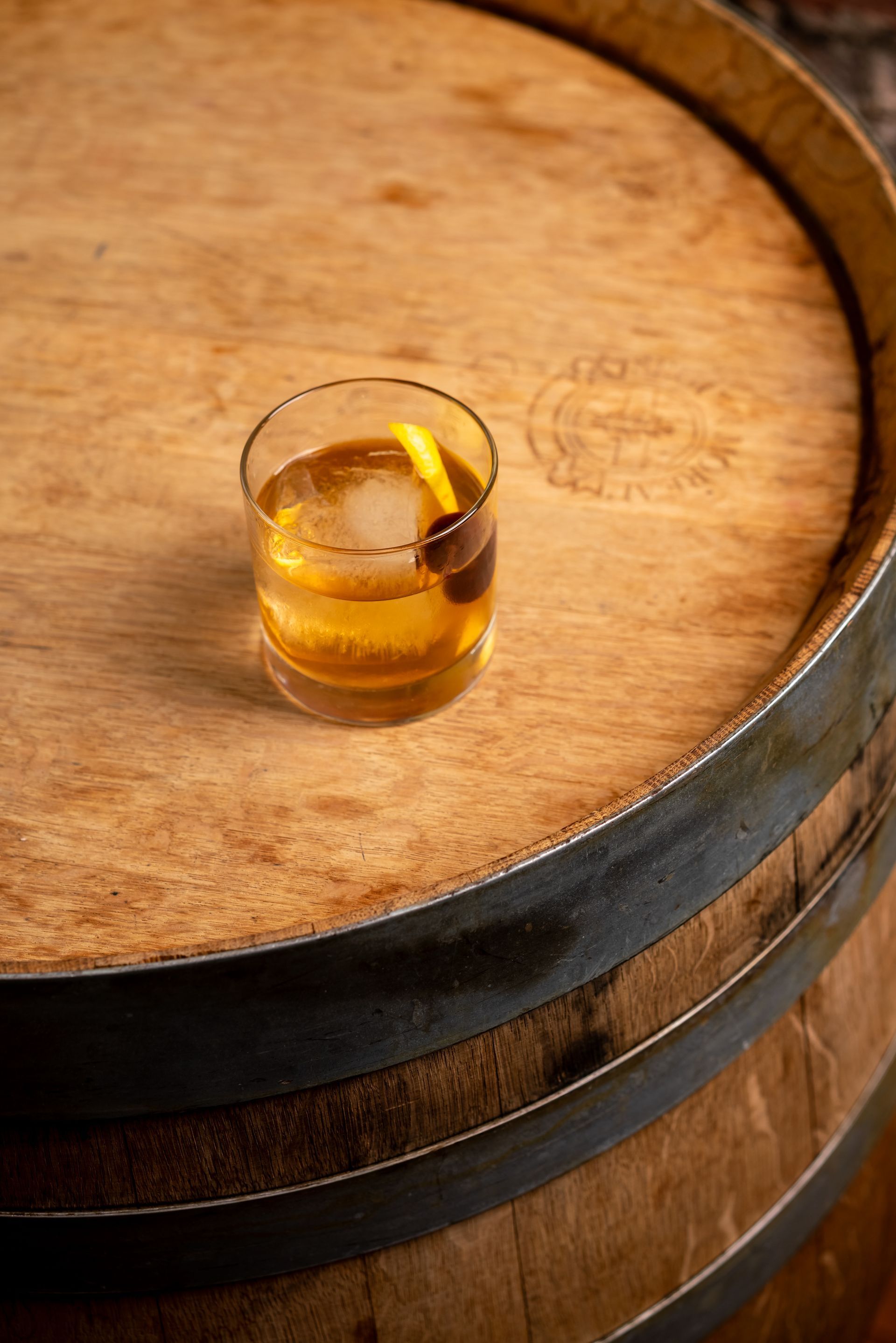 Cocktail in a rocks glass, cherry and lemon peel garnish, on a wooden barrel.