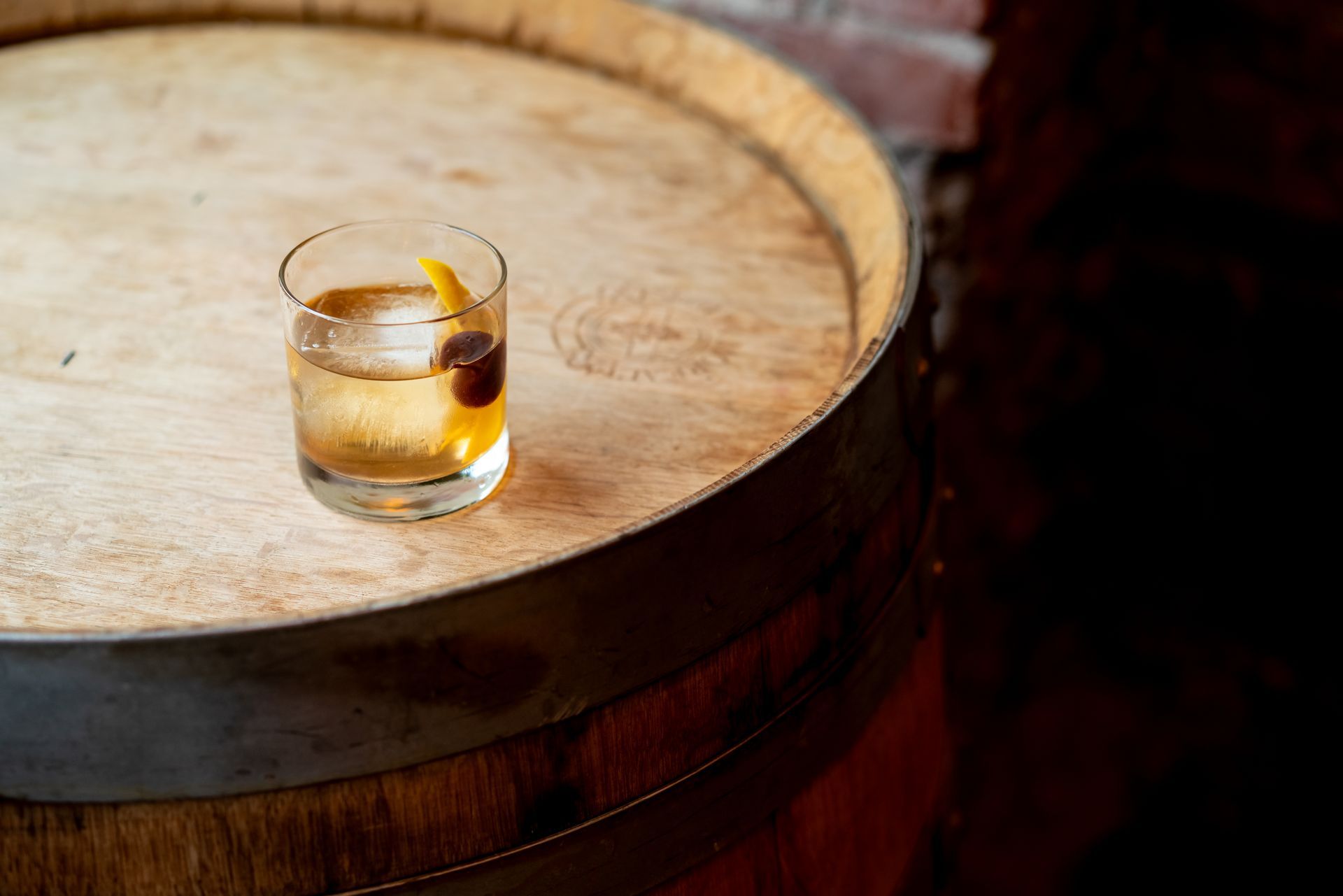 Old Fashioned cocktail on a wooden barrel, garnished with a cherry and lemon twist.