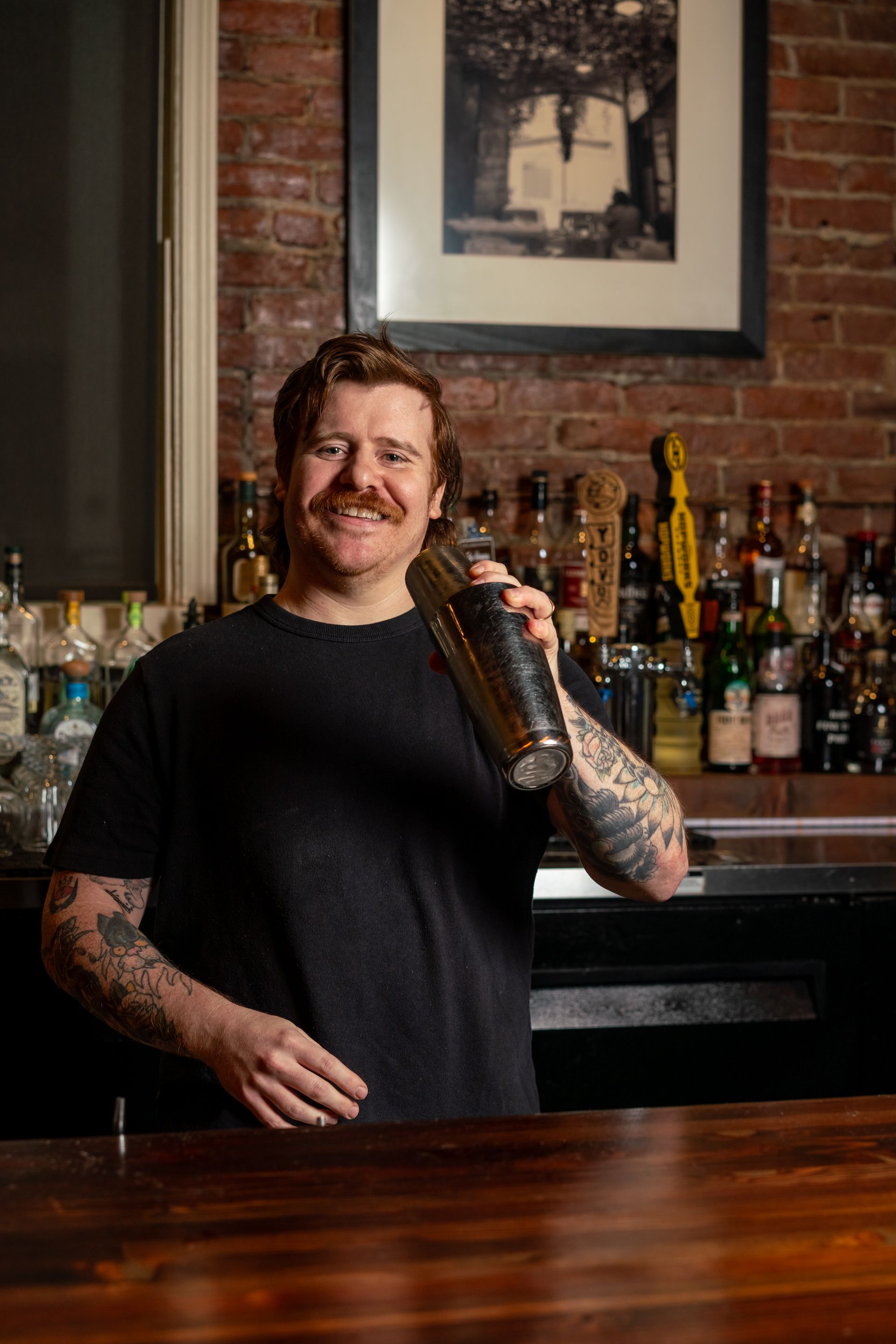 Bartender smiles while holding a cocktail shaker behind a bar, brick wall background.