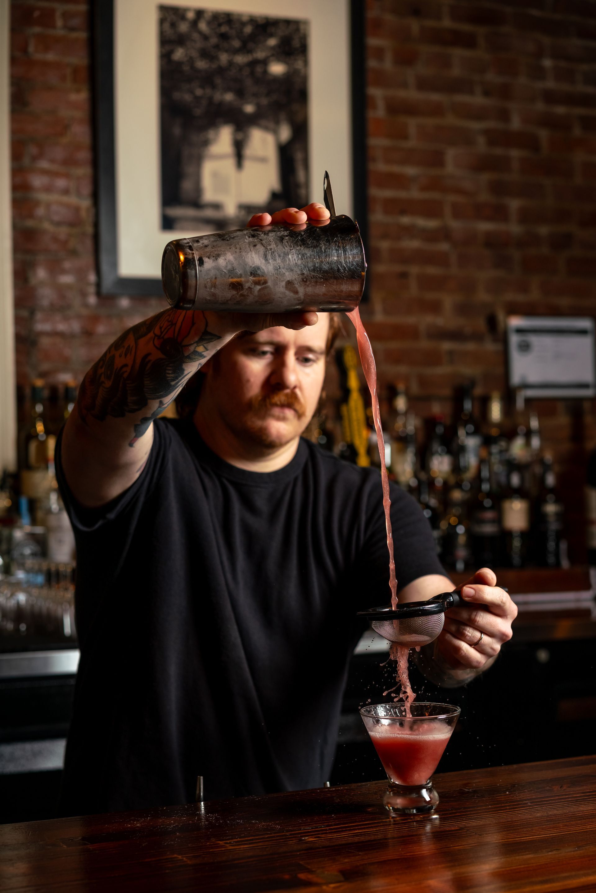 Bartender pours red cocktail from shaker into glass; bar setting.