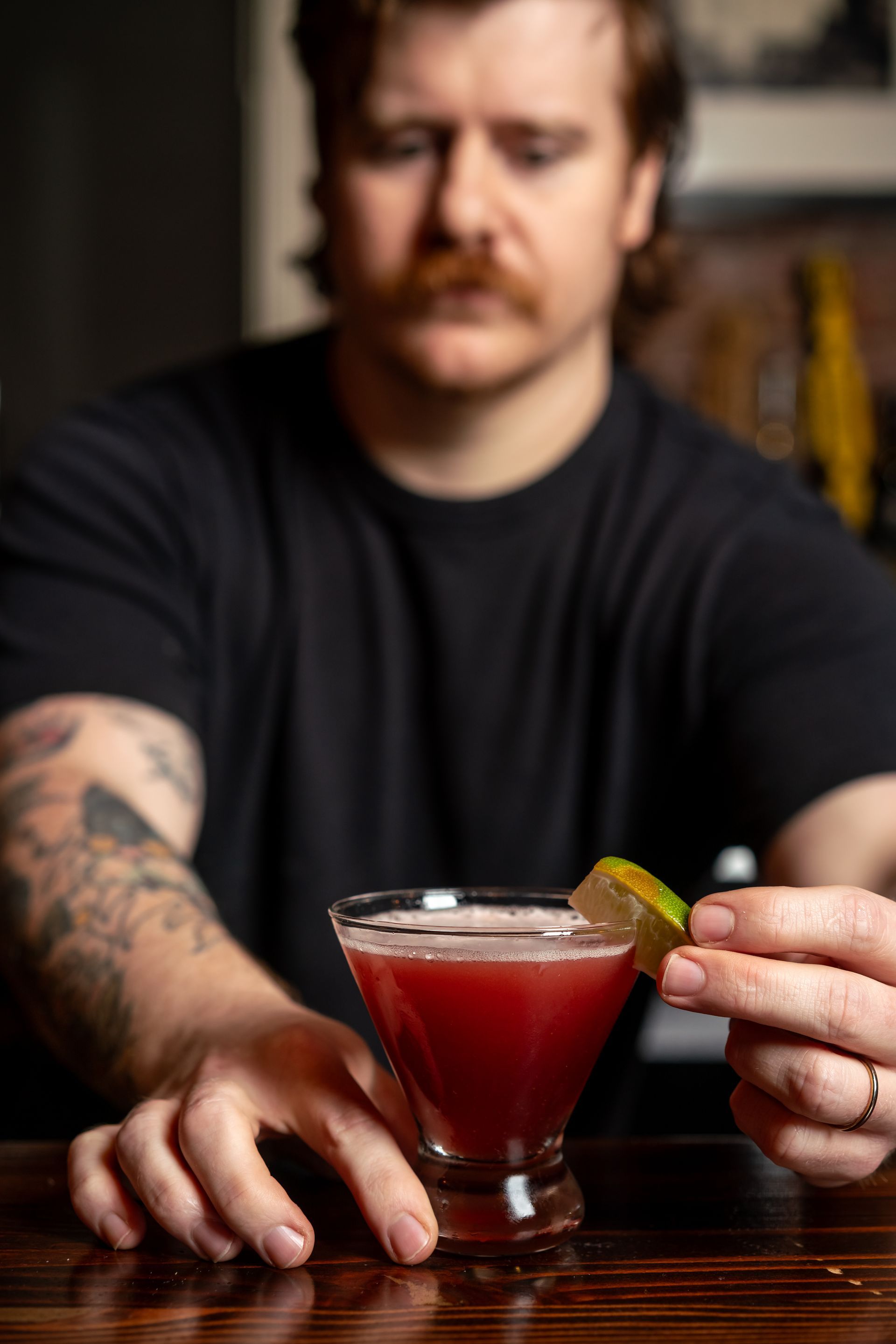 Bartender garnishes a crimson cocktail with a lime wedge, resting on a wooden bar.