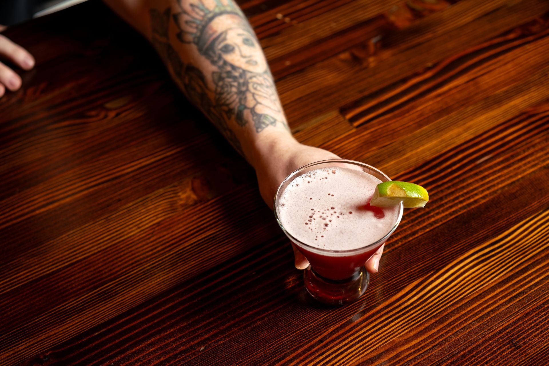 Hand holding a pink cocktail with a lime garnish on a wooden table.