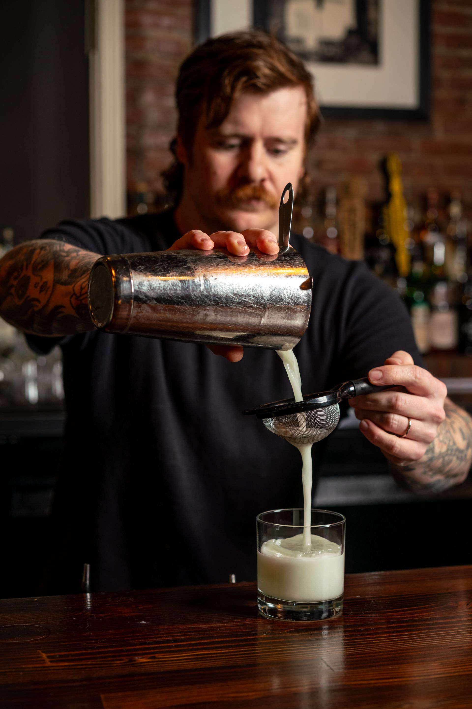 Bartender strains a light green cocktail from the shaker into a rocks glass. Bar interior, brick wall background.