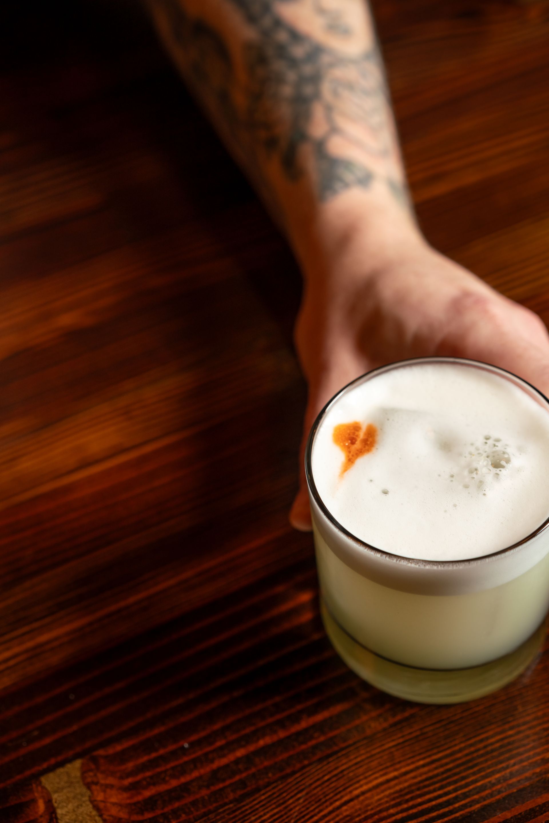 Hand holding a cocktail in a glass with a foamy top, on a dark wooden surface.