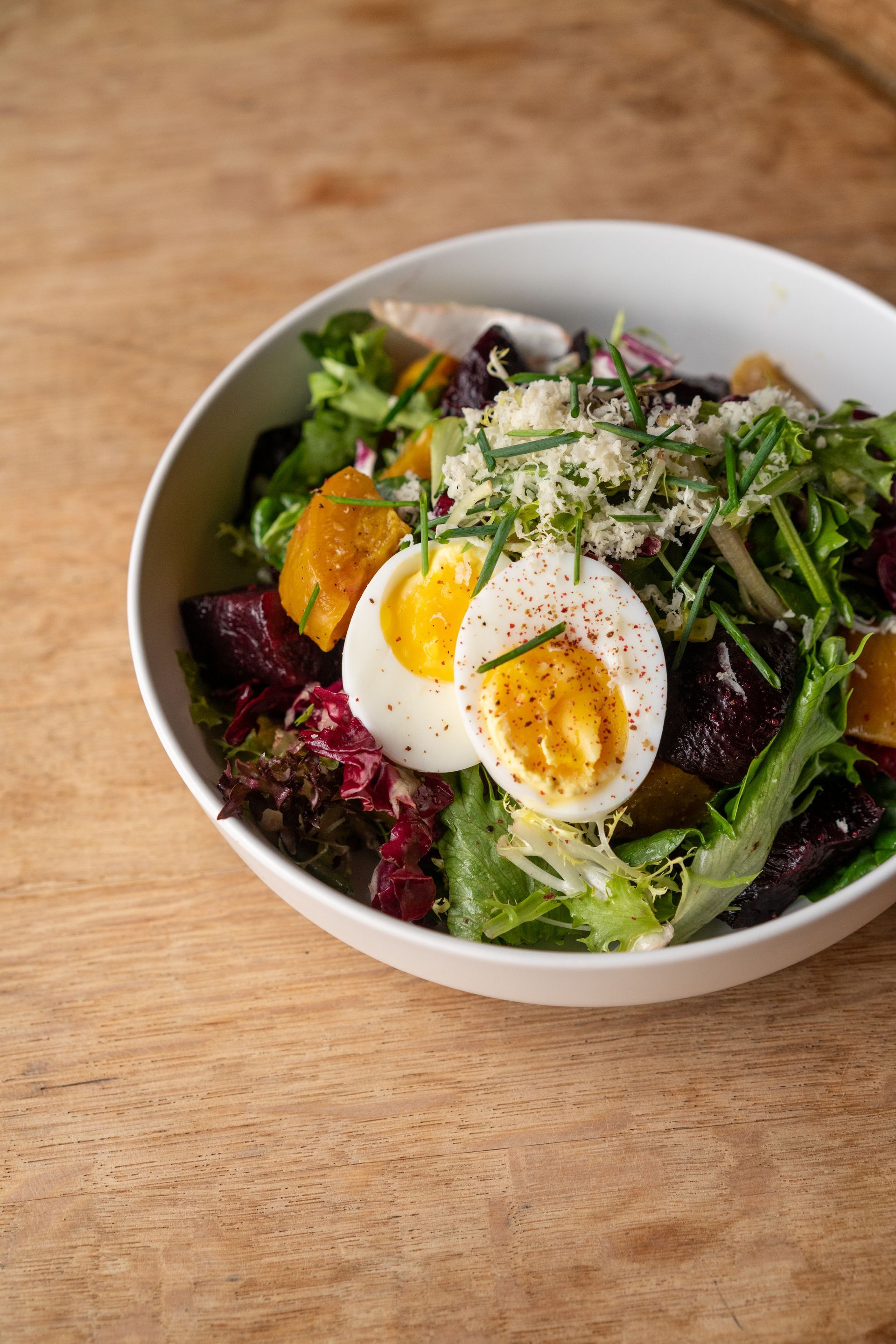 Salad in a white bowl, hard-boiled egg, beets, greens, cheese, chives, on wood table.