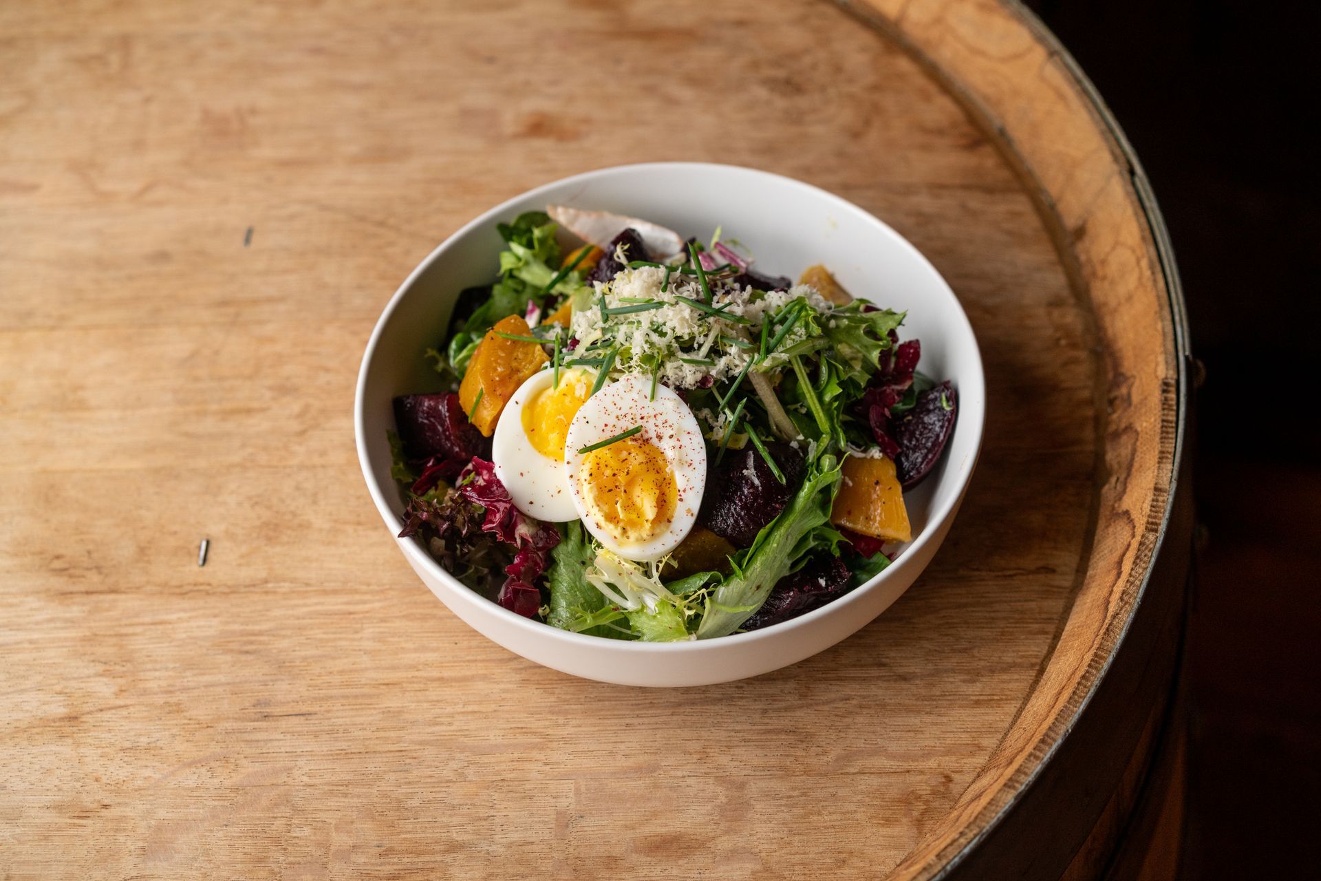 Salad with sliced hard-boiled egg, beets, greens, and cheese in a white bowl on a wooden surface.