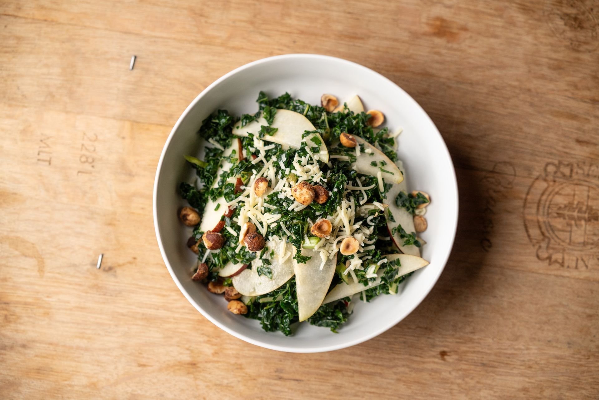 Salad in a white bowl, kale, pear slices, toasted hazelnuts, and shaved cheese on a wooden surface.