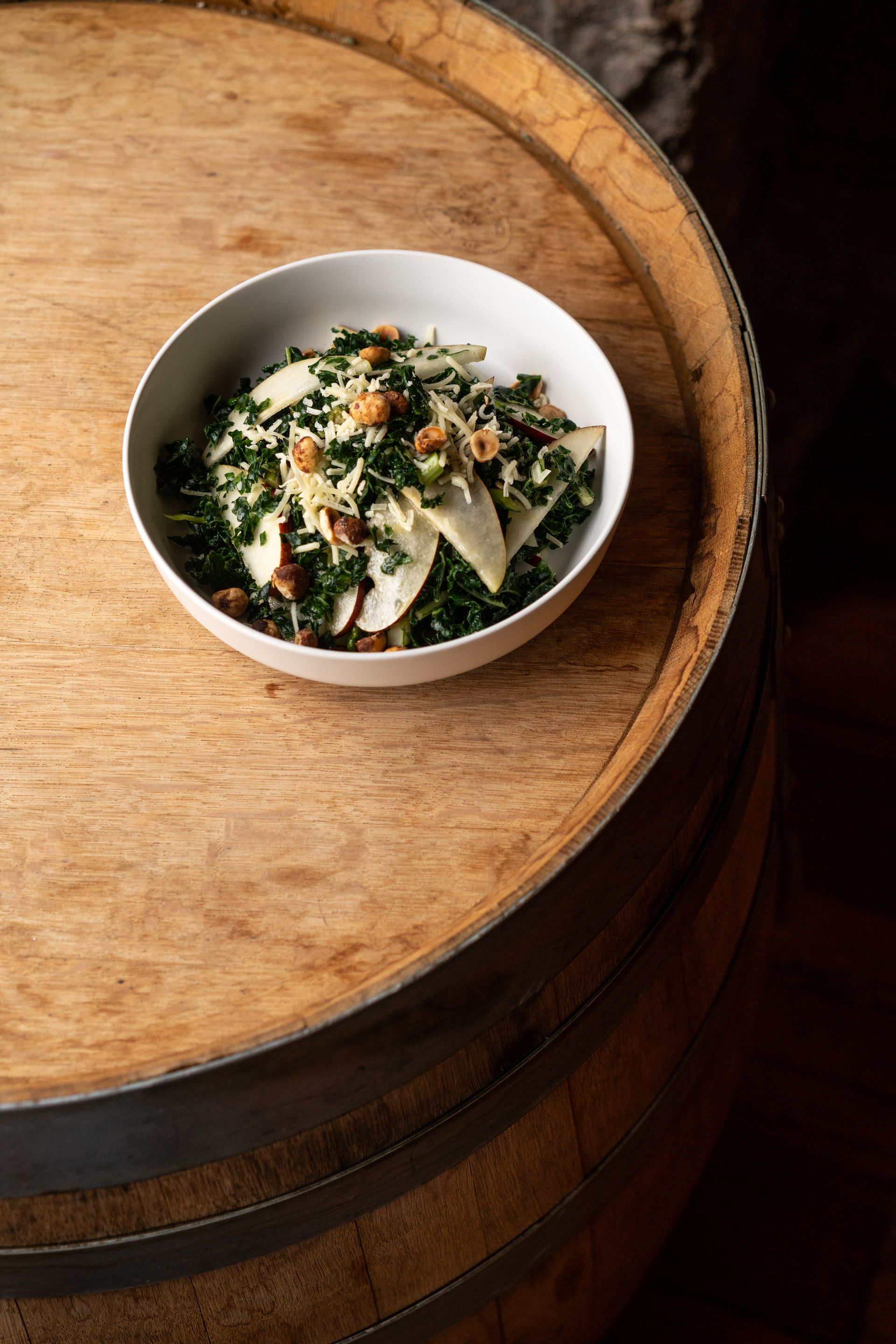 Salad in a white bowl on a wooden wine barrel, with dark leafy greens and light-colored slices.