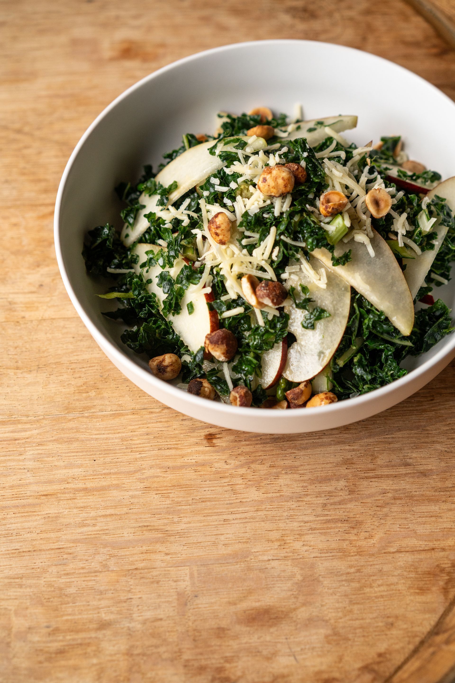 Kale salad with pear slices, hazelnuts, and grated cheese in a white bowl on wood.