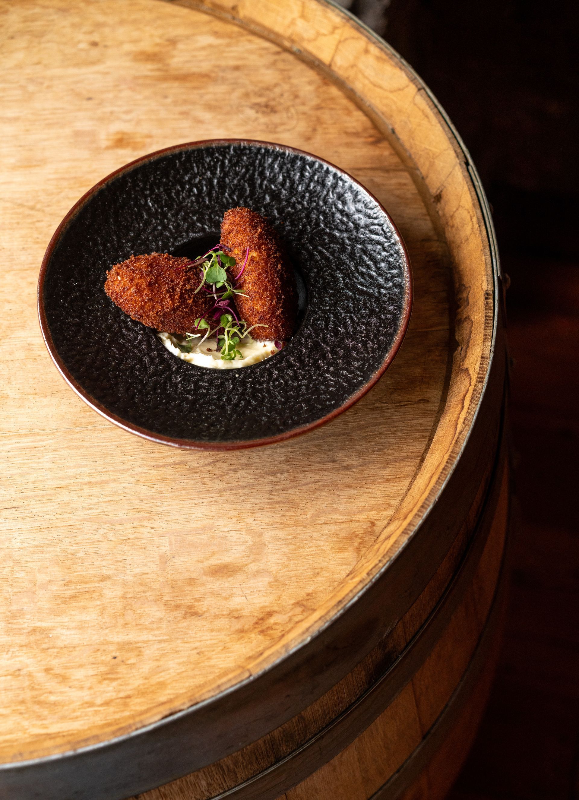 Two breaded appetizers on a black plate with a white sauce, placed on a wooden barrel.