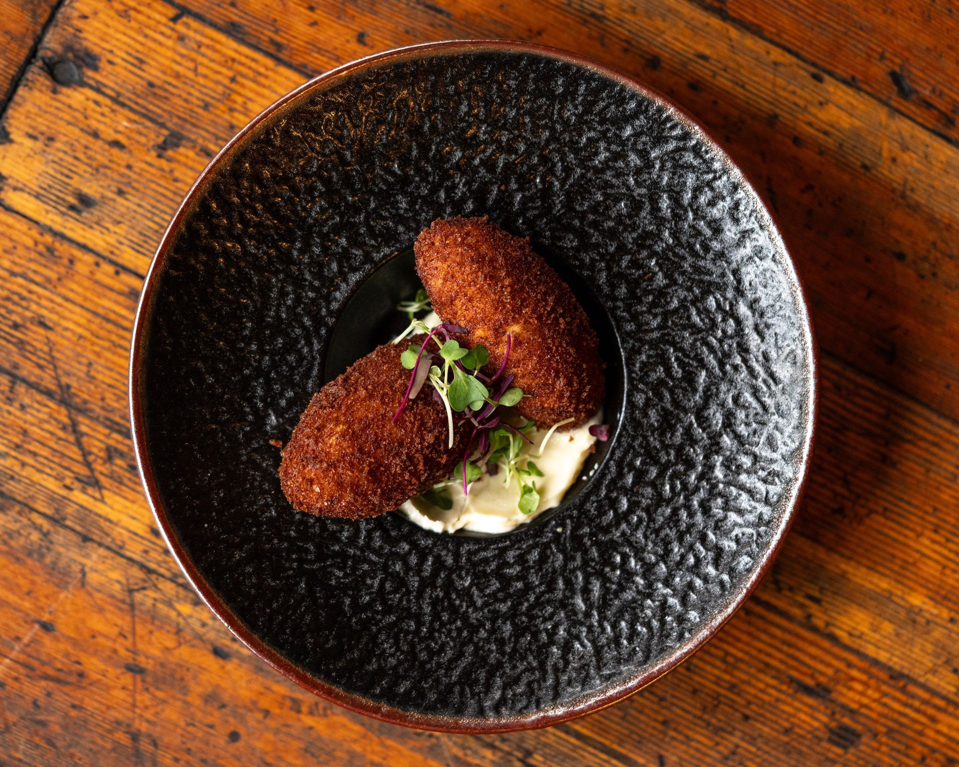 Two breaded, oblong croquettes on white sauce, garnished with sprouts, on a black textured plate.