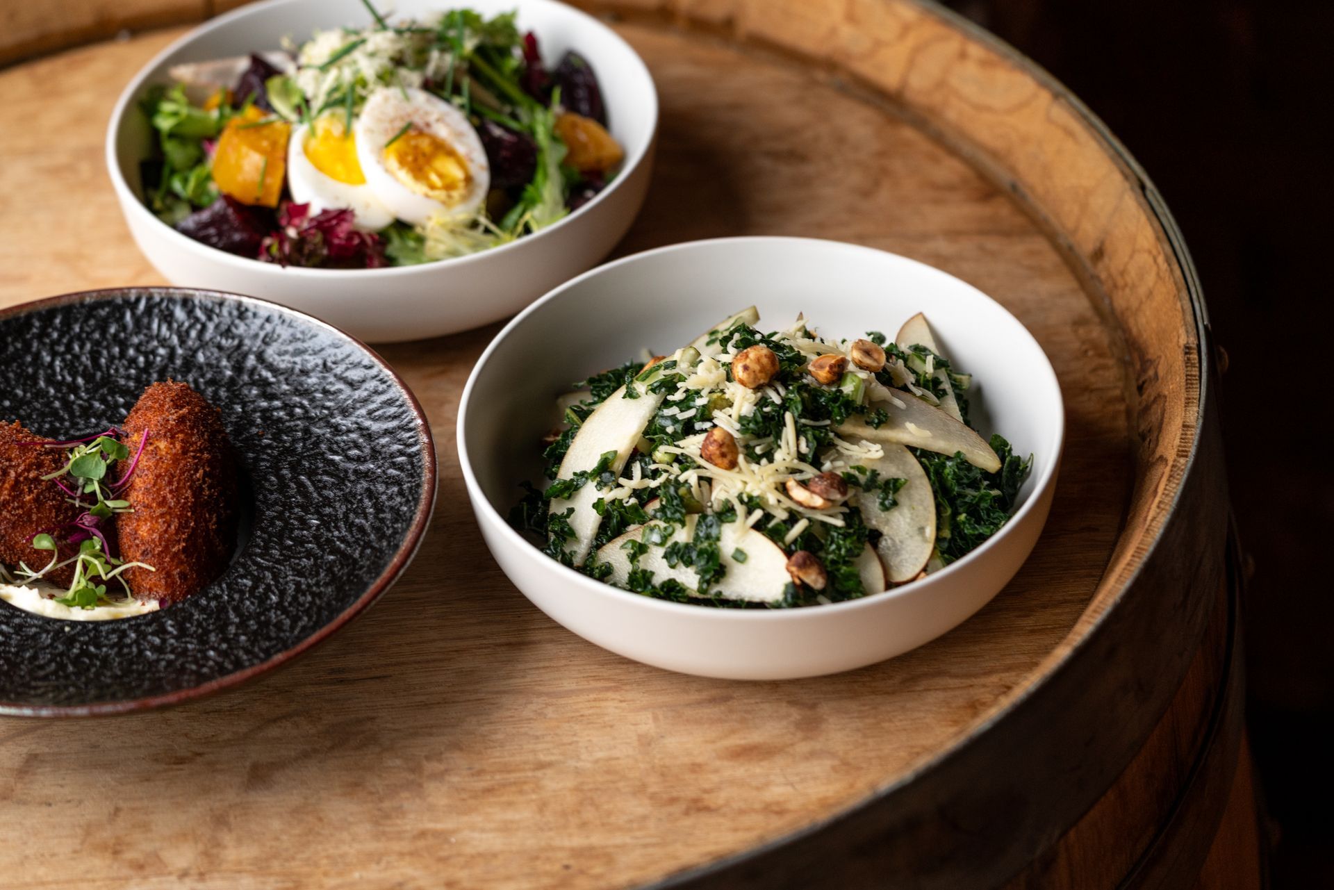 Three food dishes on a wooden barrel: salad with egg, kale salad, and fried croquettes on a black plate.