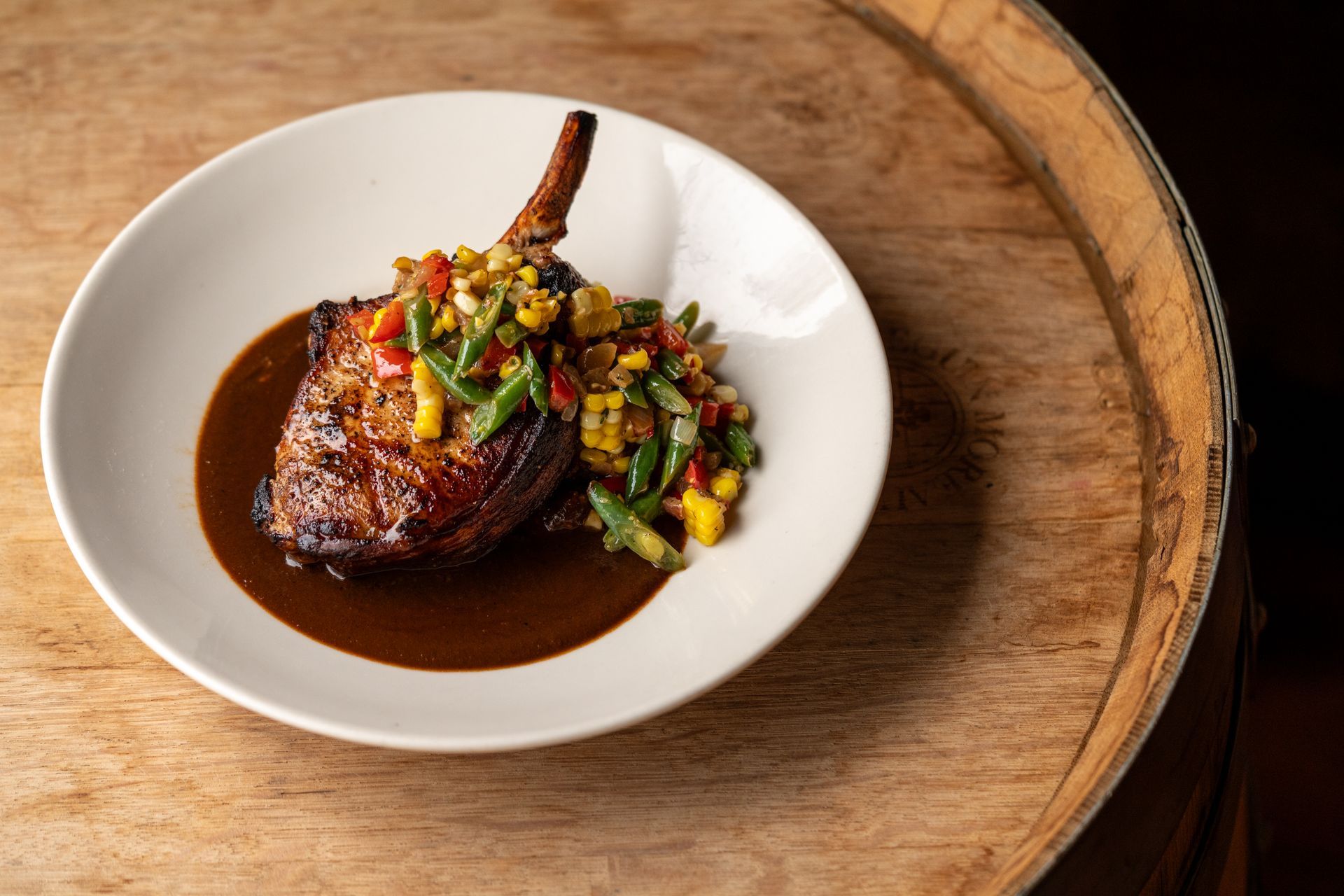 Pork chop with sauce and colorful vegetables on a white plate atop a wooden barrel.