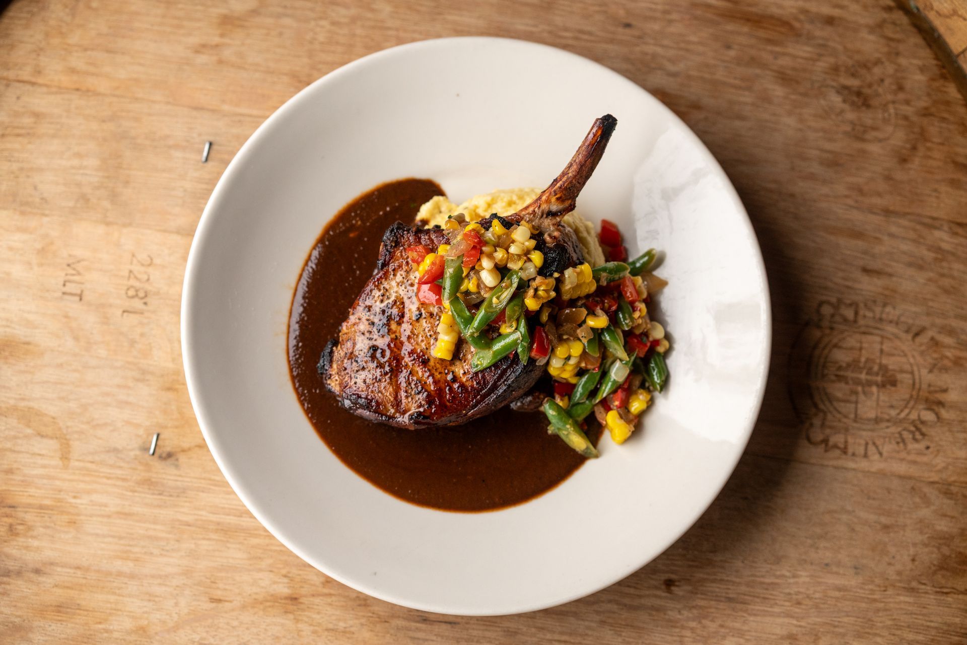Pork chop with sauce, mashed potatoes, and a vegetable medley on a white plate. Wooden table background.