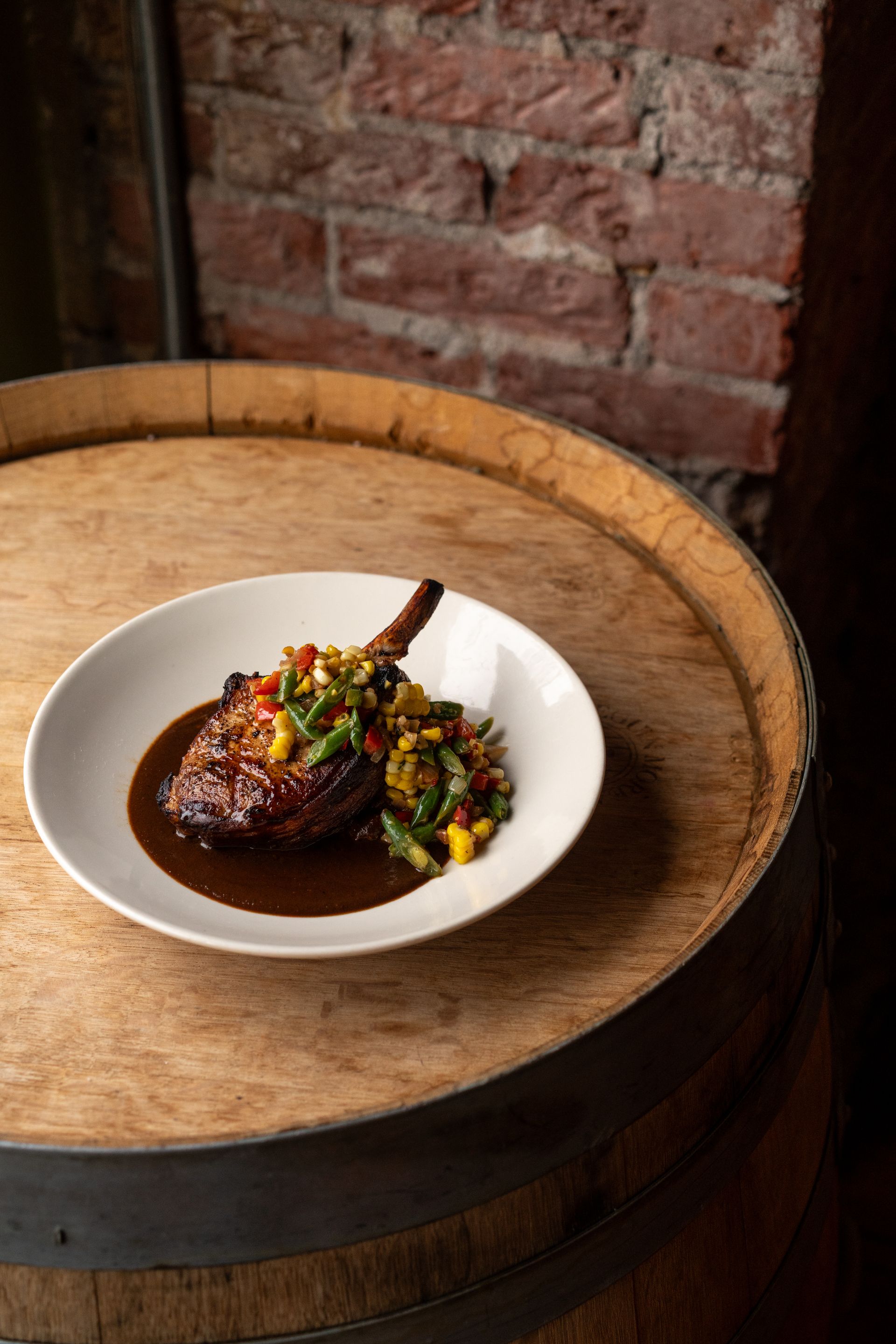 A grilled pork chop with vegetables in a sauce on a white plate, set on a wooden barrel.