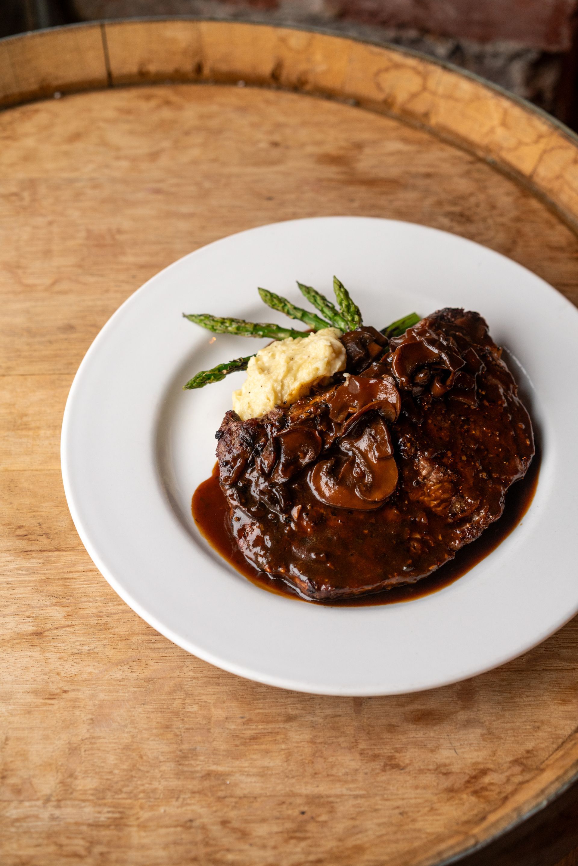 Steak with mushroom sauce, mashed potatoes, and asparagus on a white plate atop a wooden barrel.