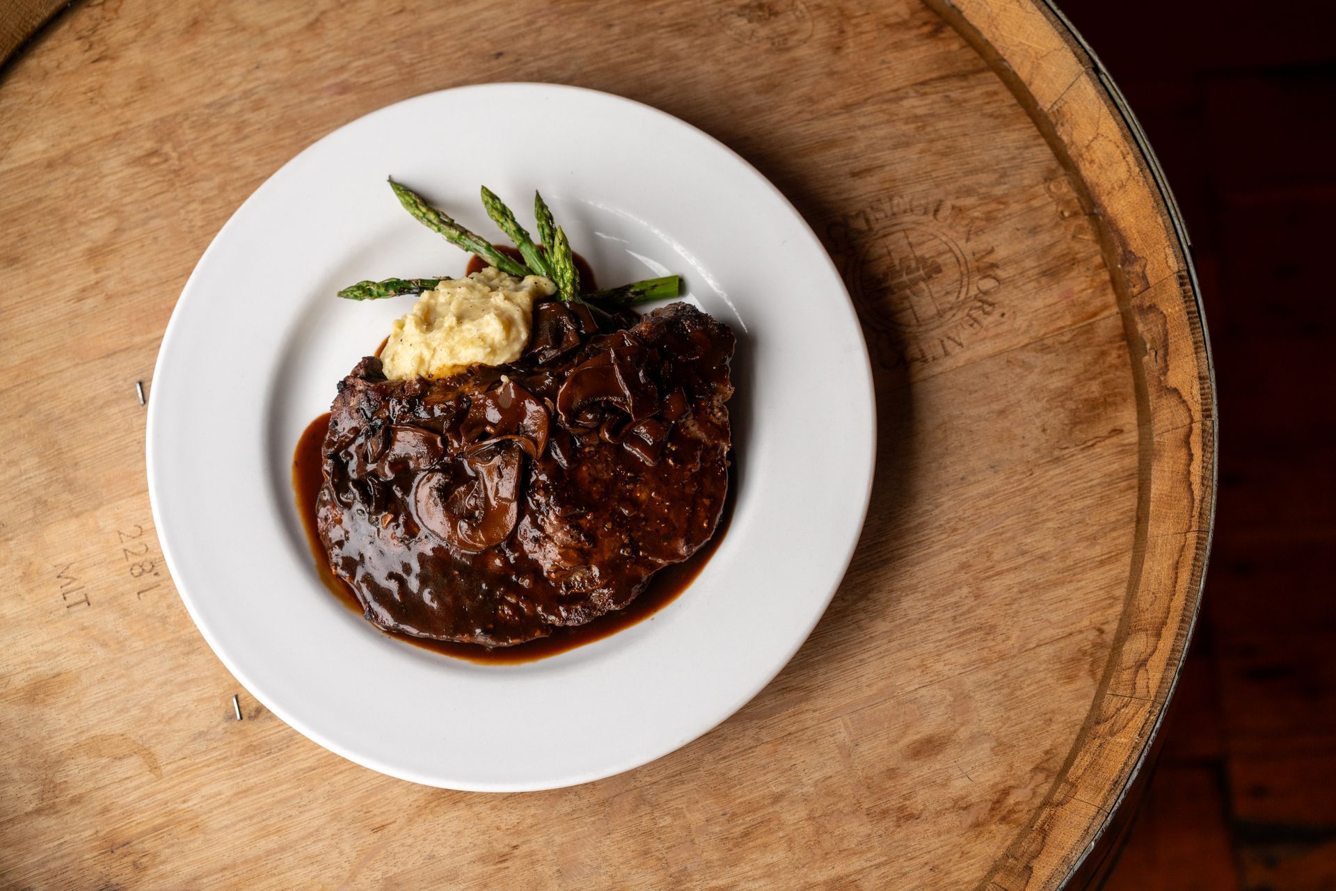 Steak with sauce and mashed potatoes on a white plate atop a wood barrel. Asparagus garnish.