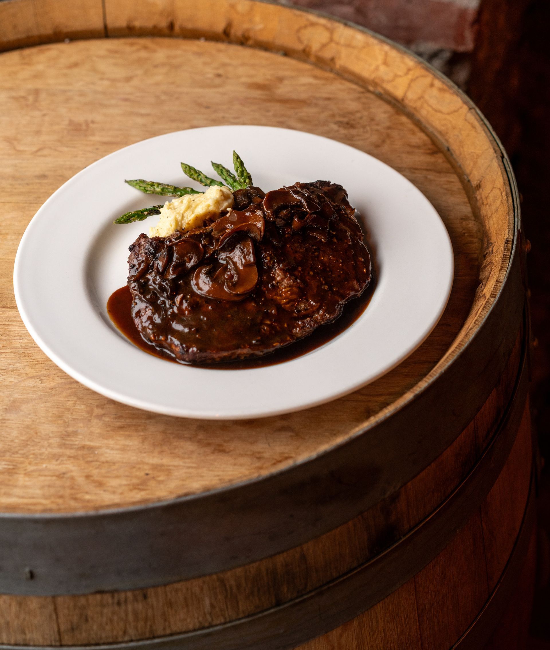 Steak with sauce, mashed potatoes, and asparagus on a white plate atop a wooden barrel.