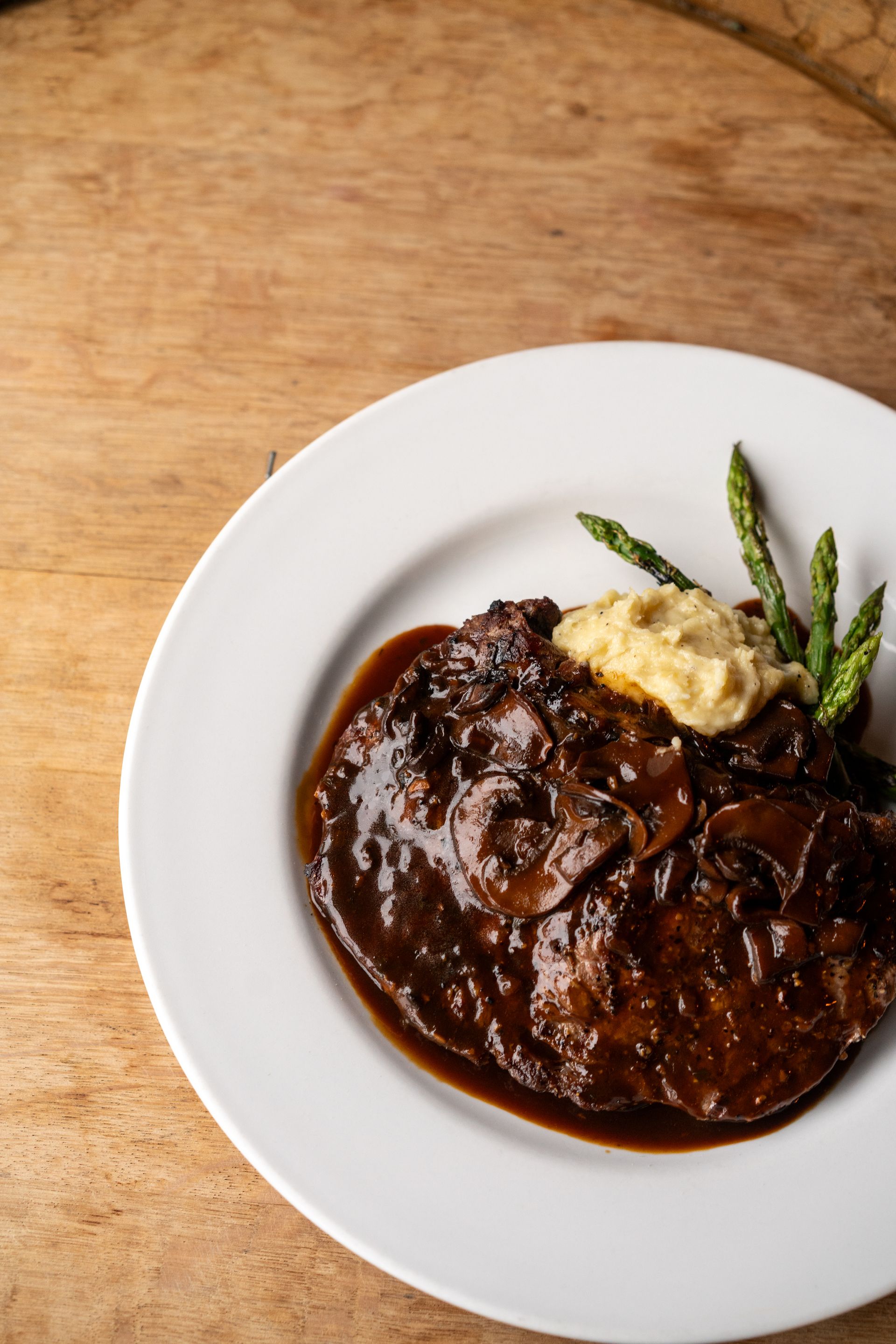 Steak with mushroom sauce, asparagus, and mashed potatoes on a white plate, wooden surface.