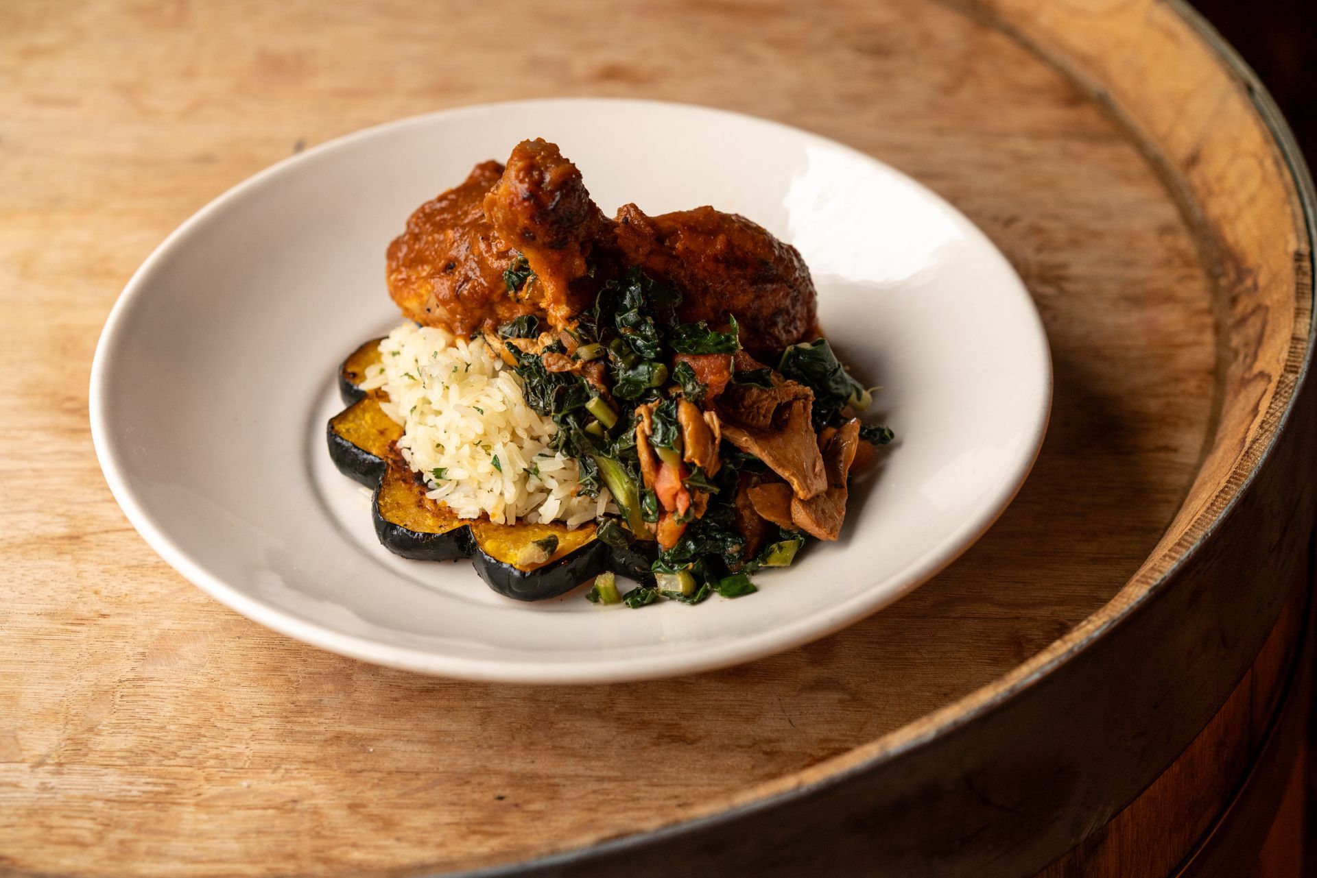 A white plate of food on a wooden barrel. Fried chicken with greens and roasted squash.