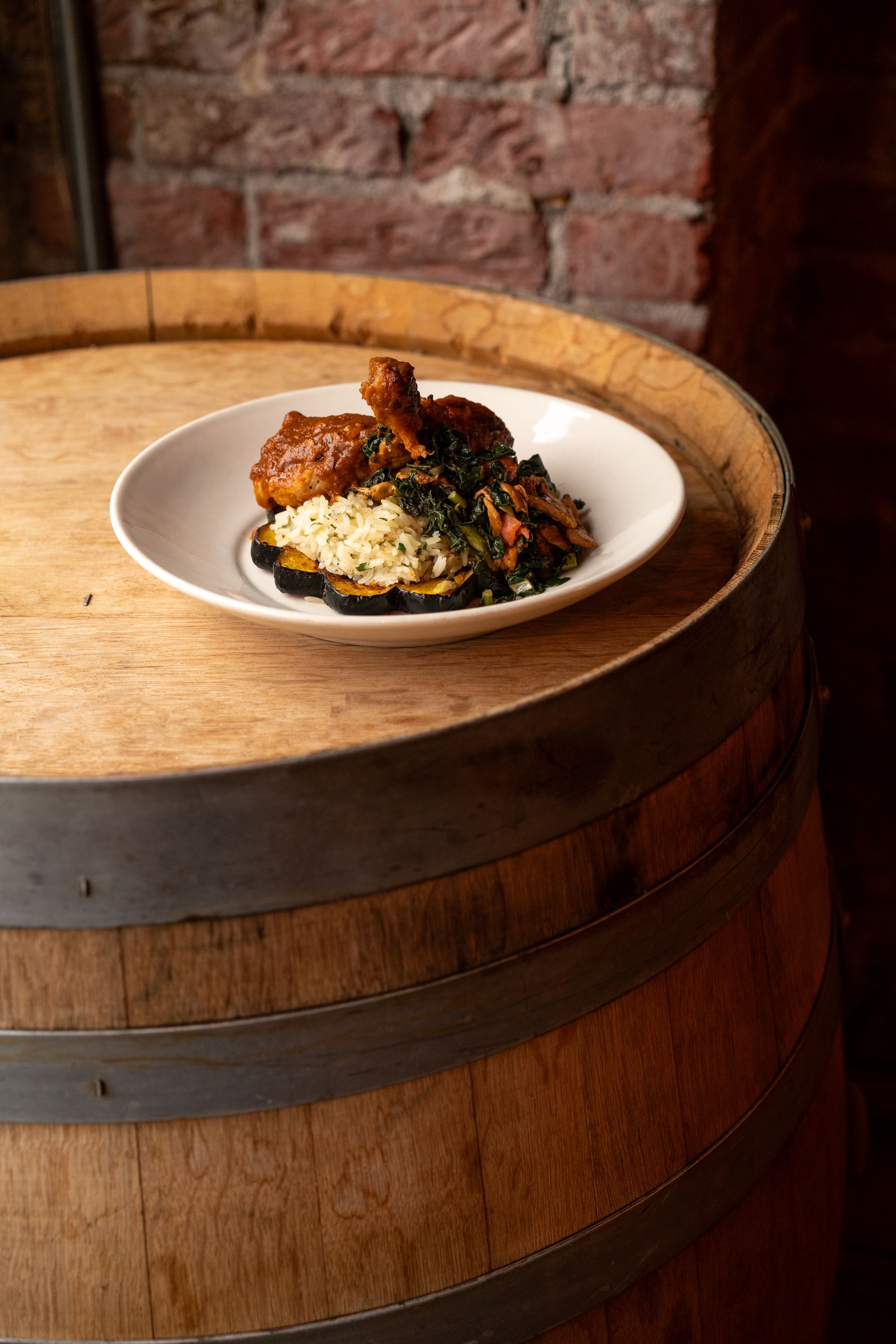 Plate of food with chicken and greens on a wooden barrel. Brick wall in the background.