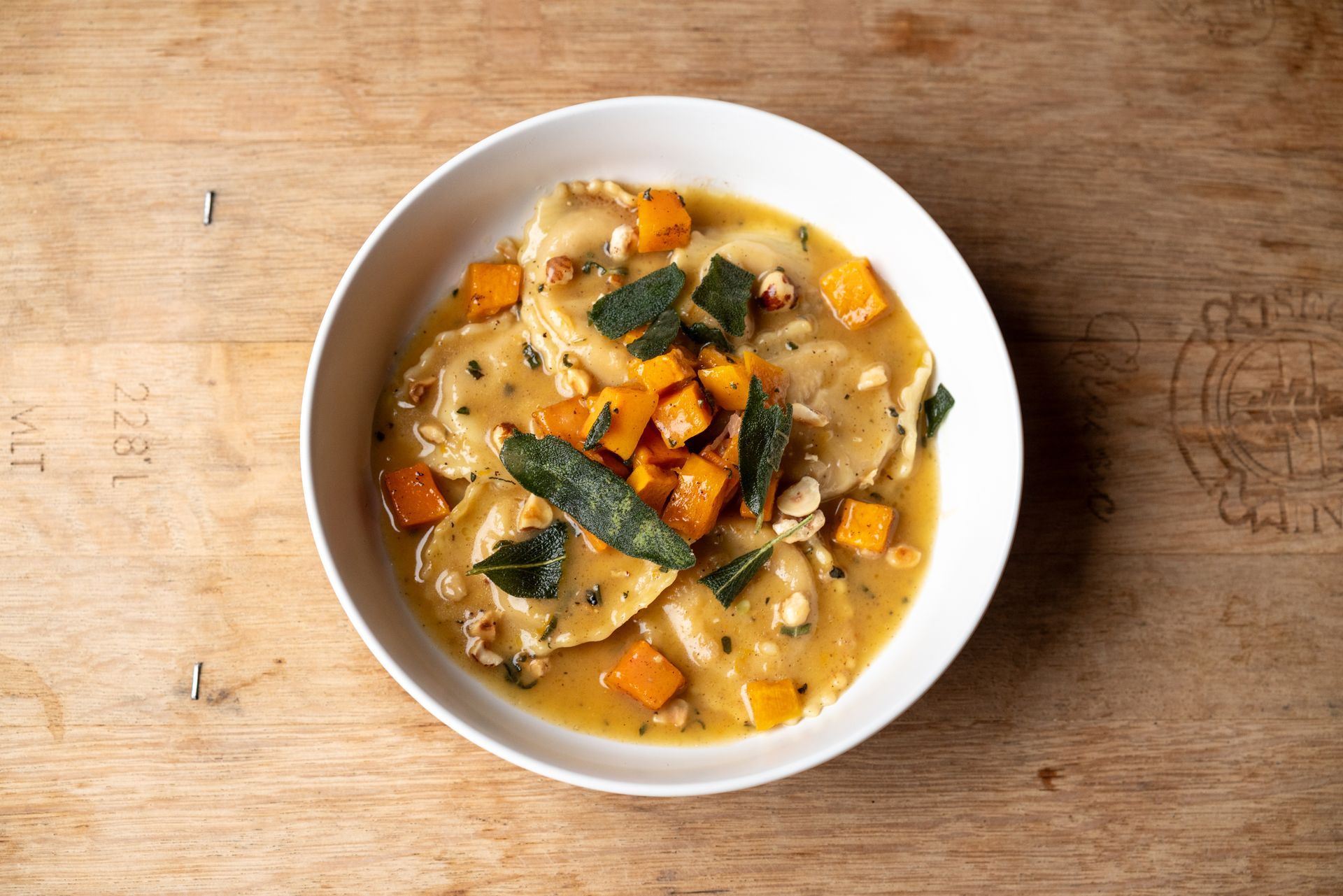 Bowl of ravioli with butternut squash, sage, and walnuts on a wooden surface.