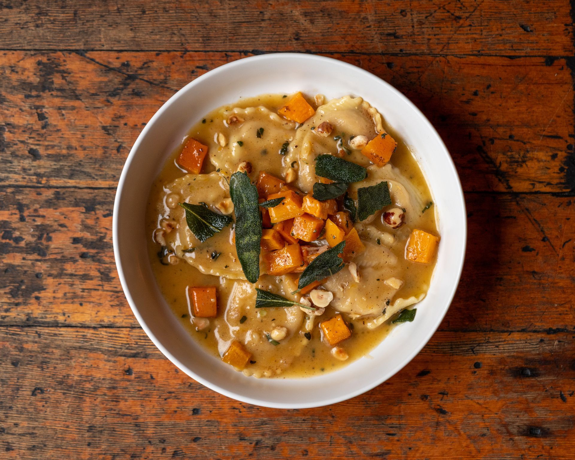 Butternut squash ravioli in a white bowl with sage, nuts, and orange cubes on a wooden table.