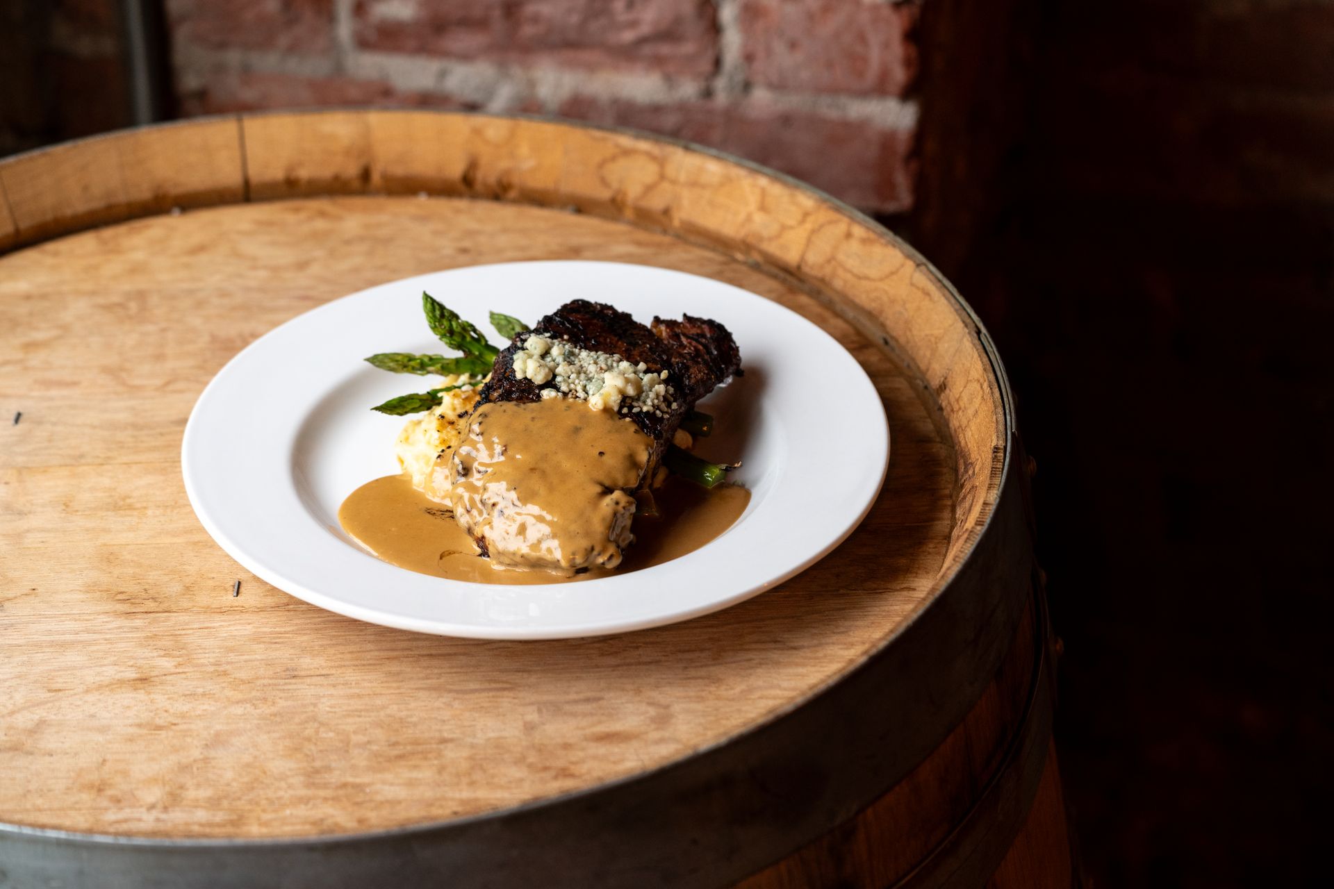 Plate of food on a wooden barrel, steak with sauce, mashed potatoes, asparagus, and a topping.