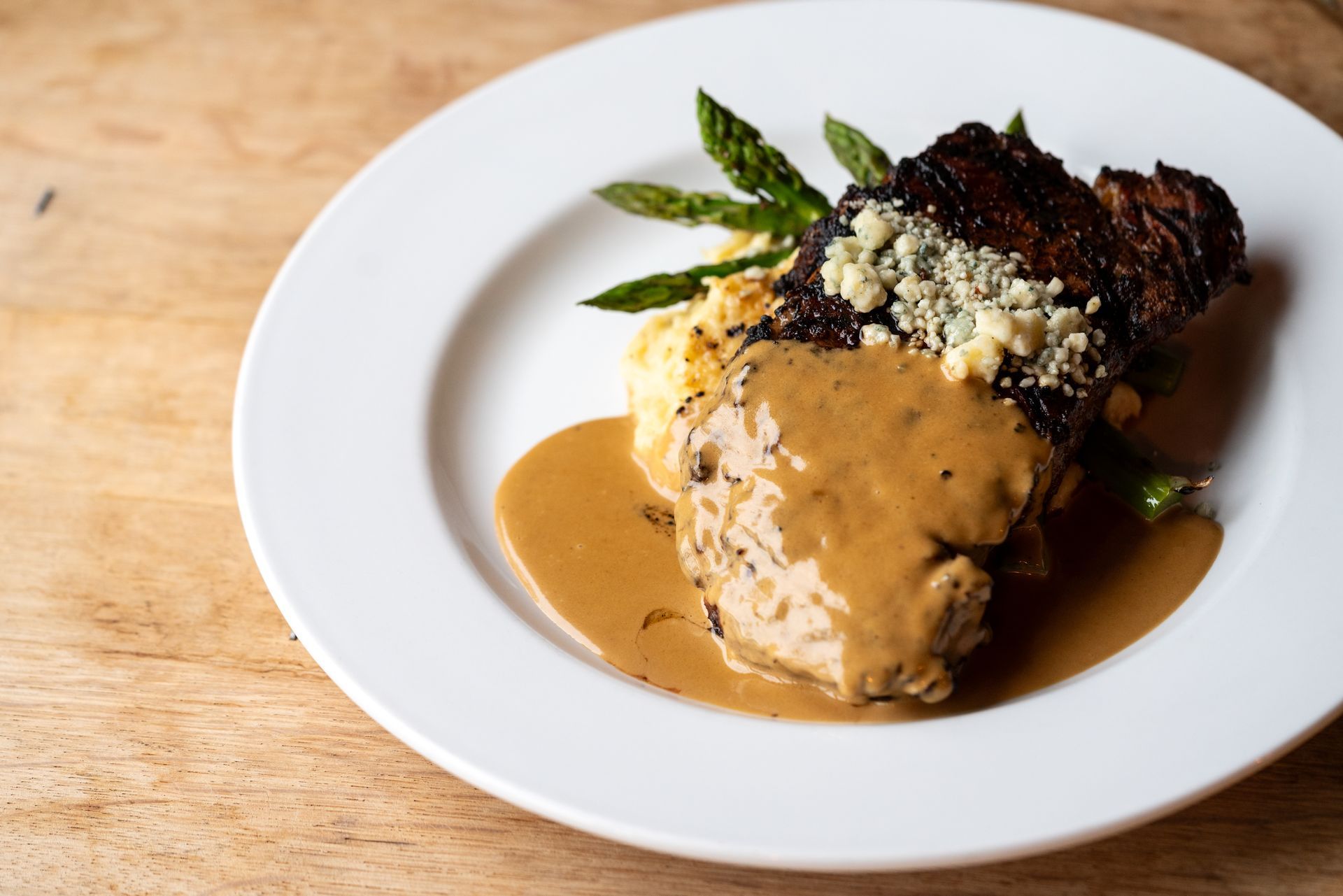 Steak with gravy, mashed potatoes, asparagus, and blue cheese on a white plate.