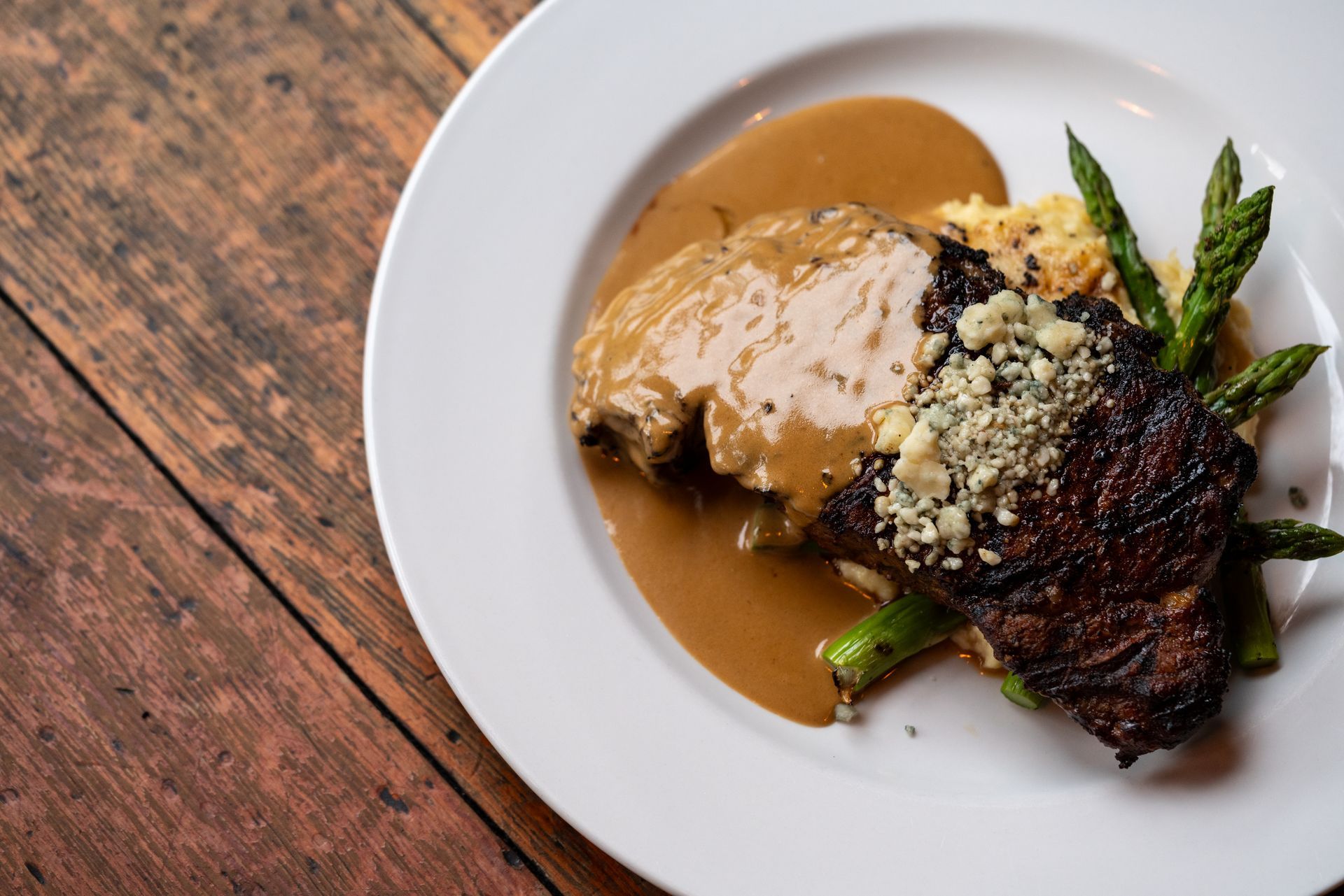 Steak with sauce, asparagus, and crumbled blue cheese on a white plate over a wooden table.
